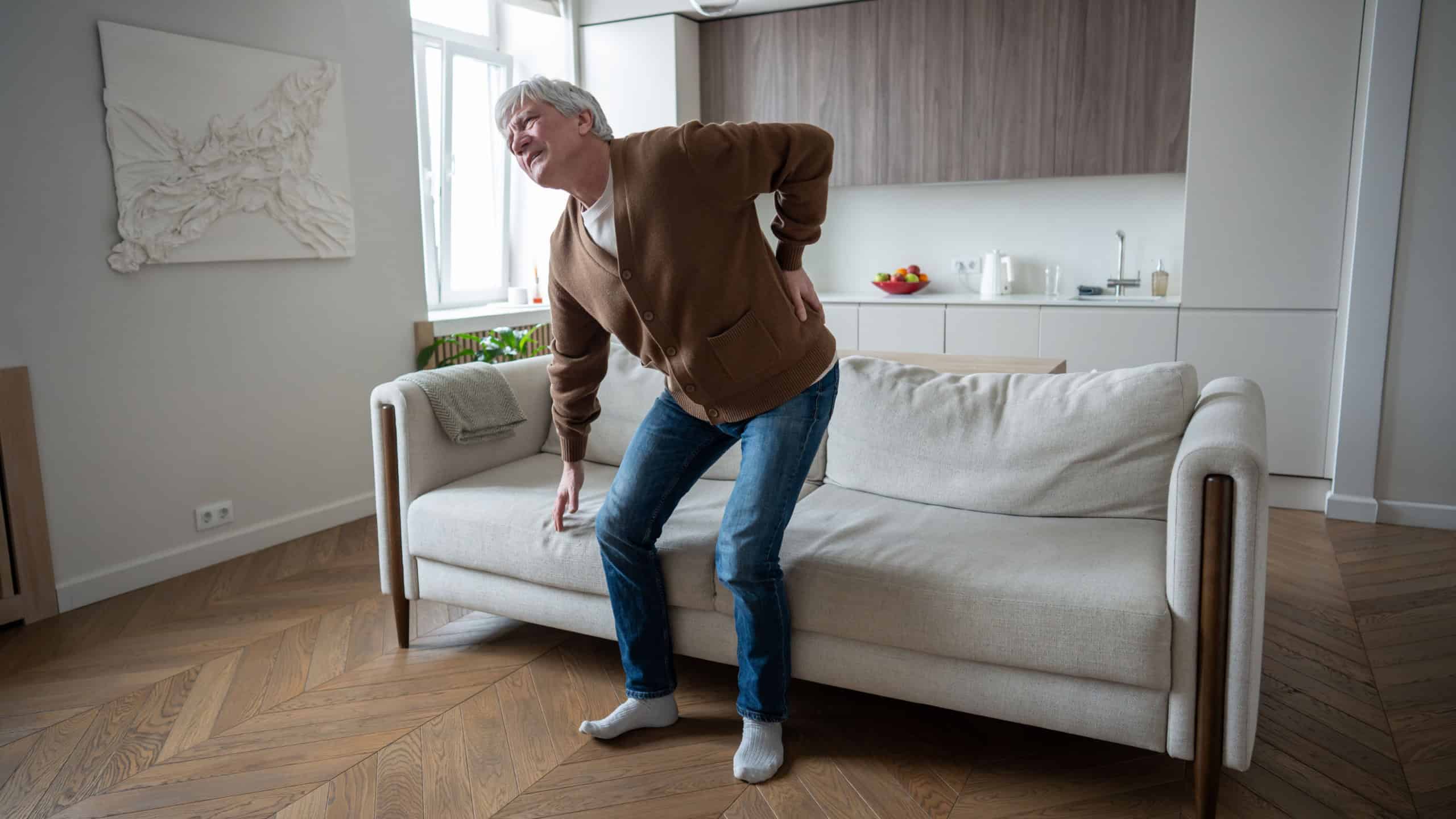 Senior man grimacing in pain attempting to stand up from couch due to spinal condition. Chronic osteochondrosis, vertebral degeneration, limited mobility, age related disorders, movement difficulties