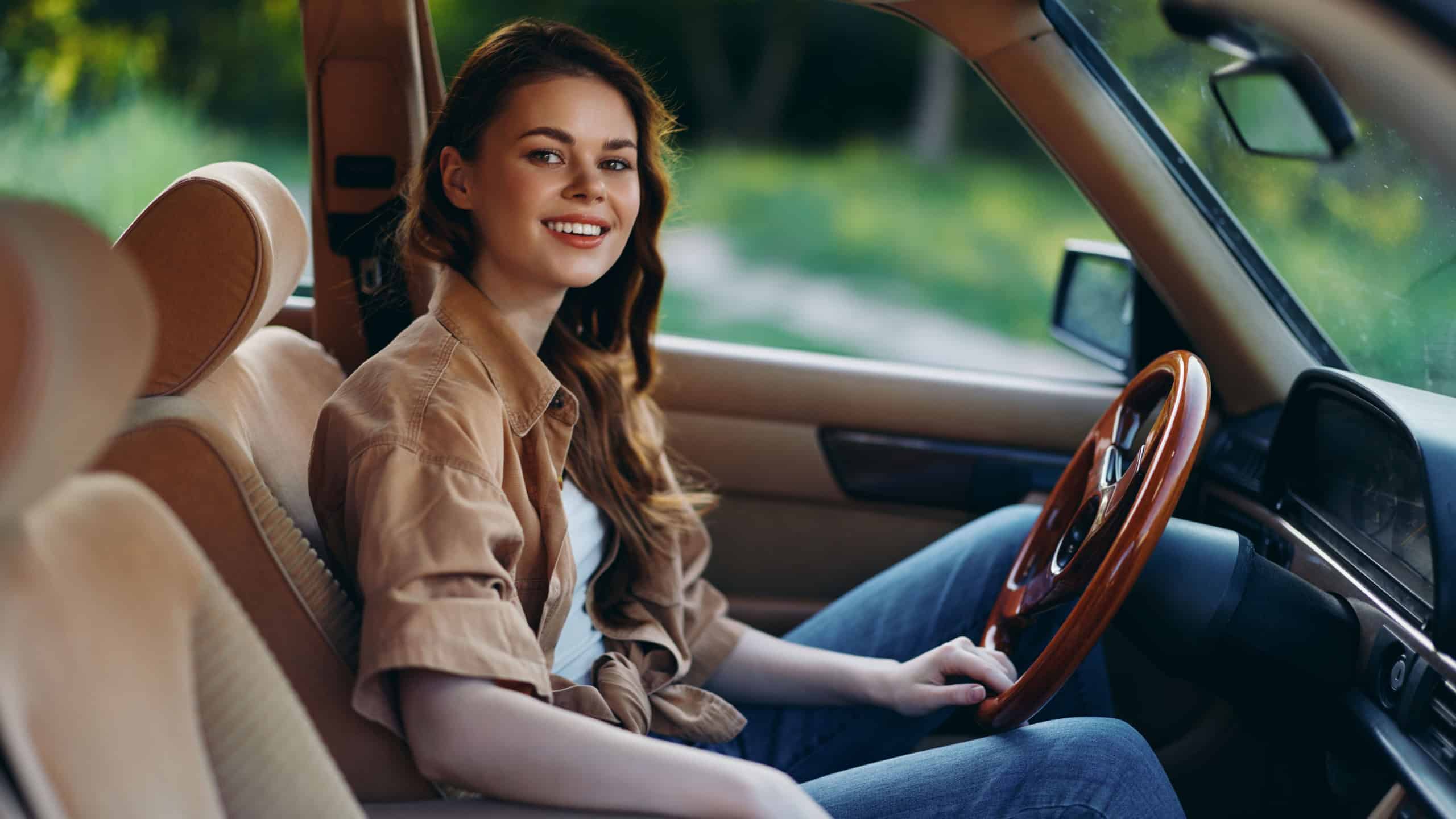 Cheerful young woman sitting in a vintage car, enjoying a summer drive in nature, wearing a casual brown shirt and smiling warmly.