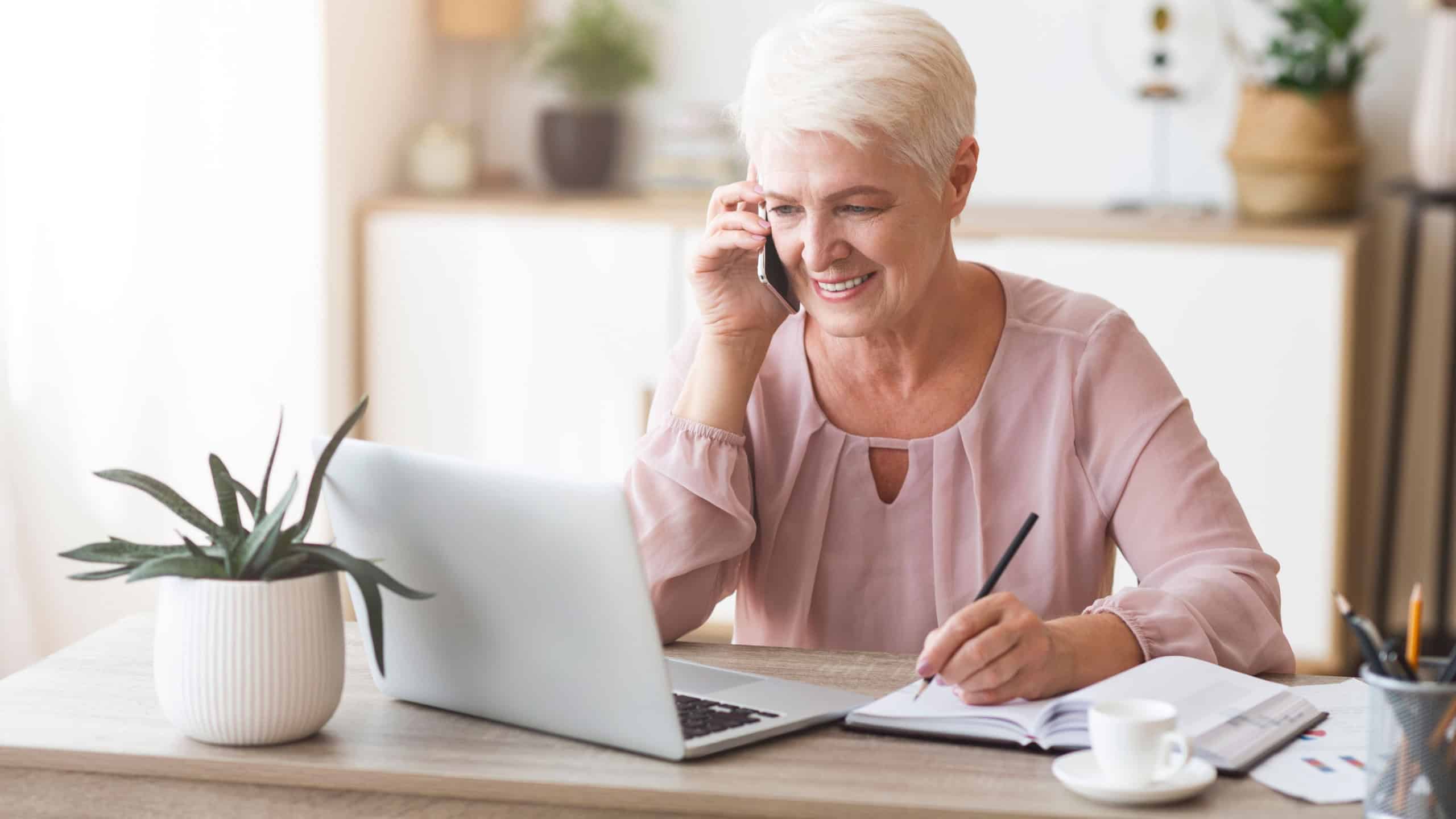 Senior business woman working from home, discussing project with client, making notes in front of laptop, panorama with empty space