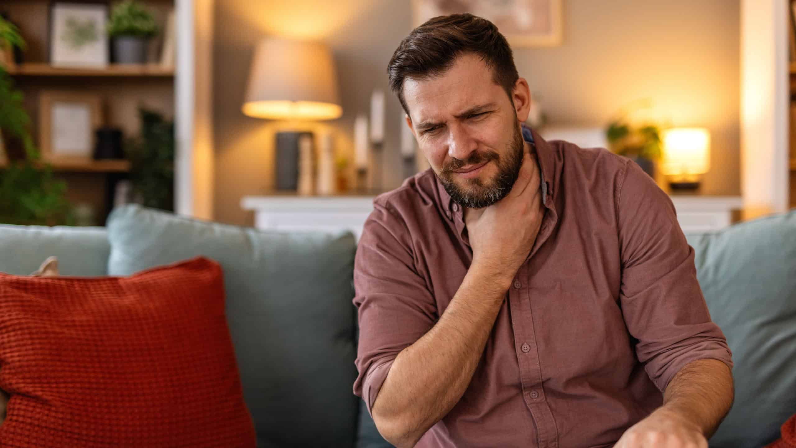 Man sitting on a sofa, holding his throat in discomfort, feeling a sore throat. He experiences a moment of pain while resting at home, reflecting the struggle of dealing with a cold or illness.