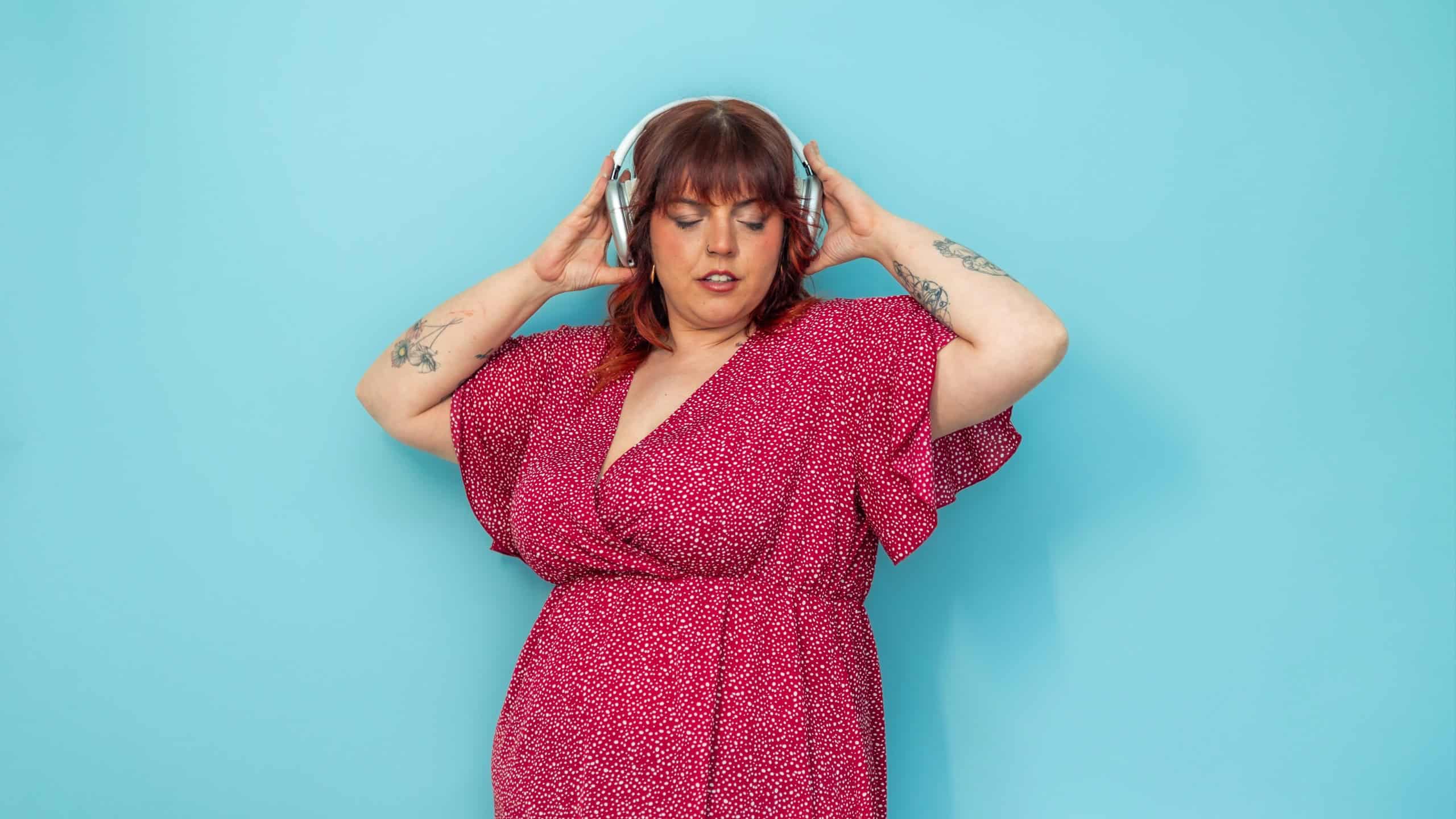 Plus-size woman with red hair and tattoos wearing a red dress, holding headphones on her ears with closed eyes, enjoying music on a vibrant blue background, conveying a sense of joy and relaxation