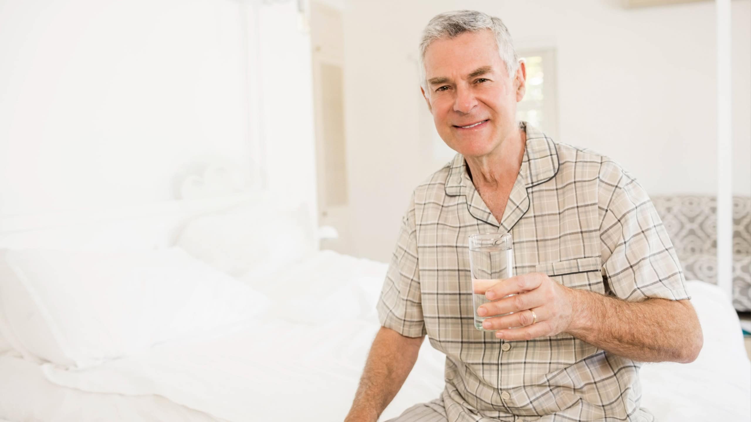Smiling senior man sitting on bed in bright bedroom, holding glass of water, copy space. Relaxation, wellness, comfort, tranquility, health, cozy, residential
