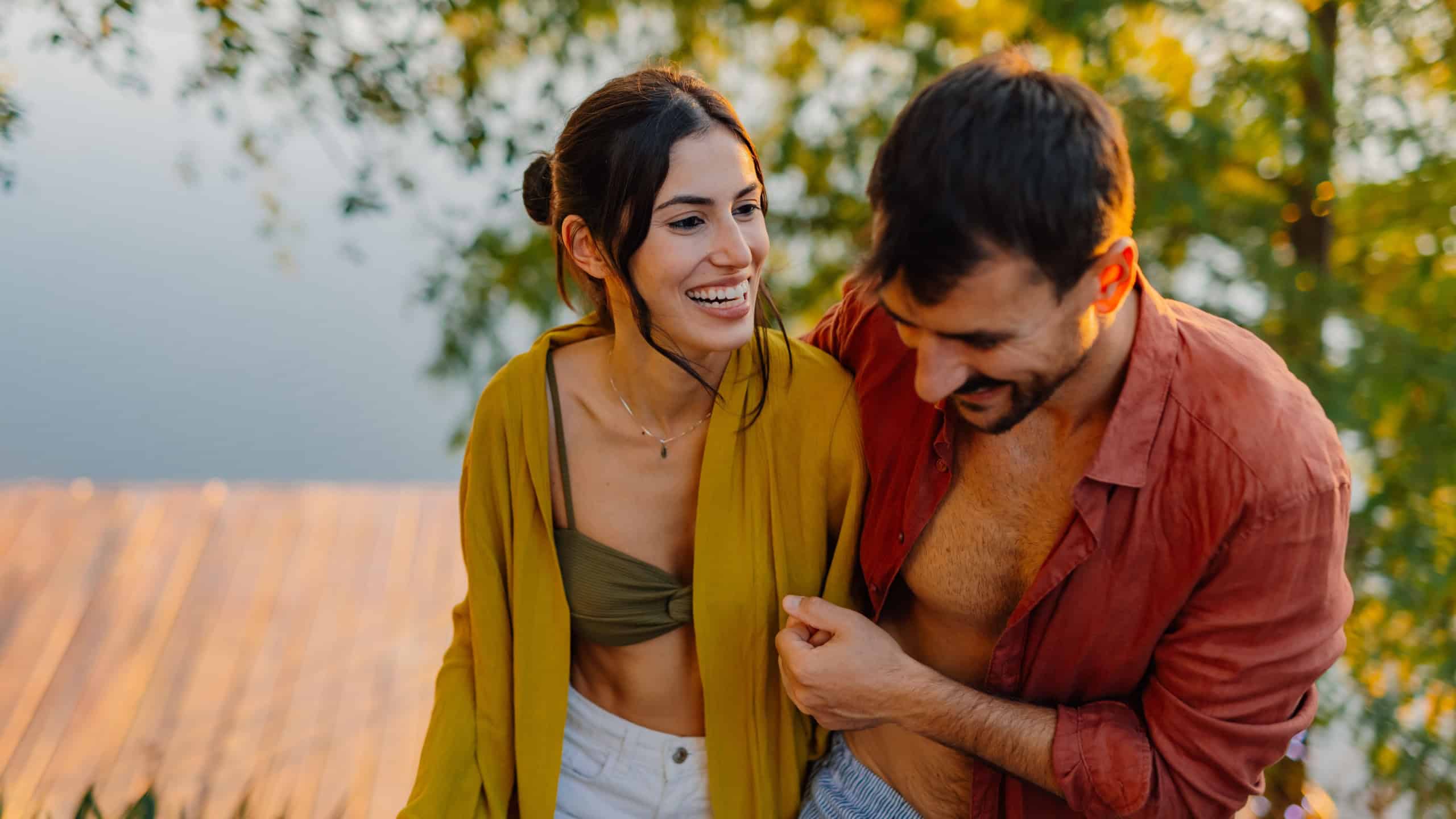 Young couple laughing and embracing near a serene lake at sunset, cherishing a romantic moment together while enjoying their vacation in a picturesque outdoor setting