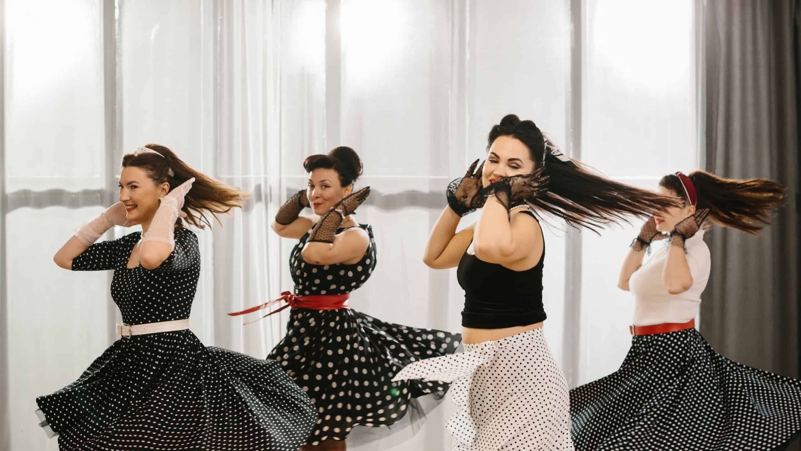Four female dancers wearing polka dot dresses and skirts are dancing a swing dance in a bright room with a 1950s style