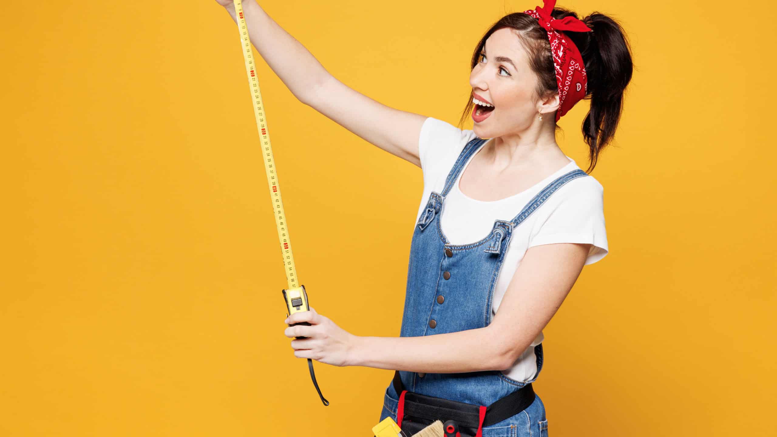 Young surprised employee laborer handyman woman wears overalls belt hold tape measure isolated on plain yellow background. Instruments accessories for renovation apartment room. Repair home concept
