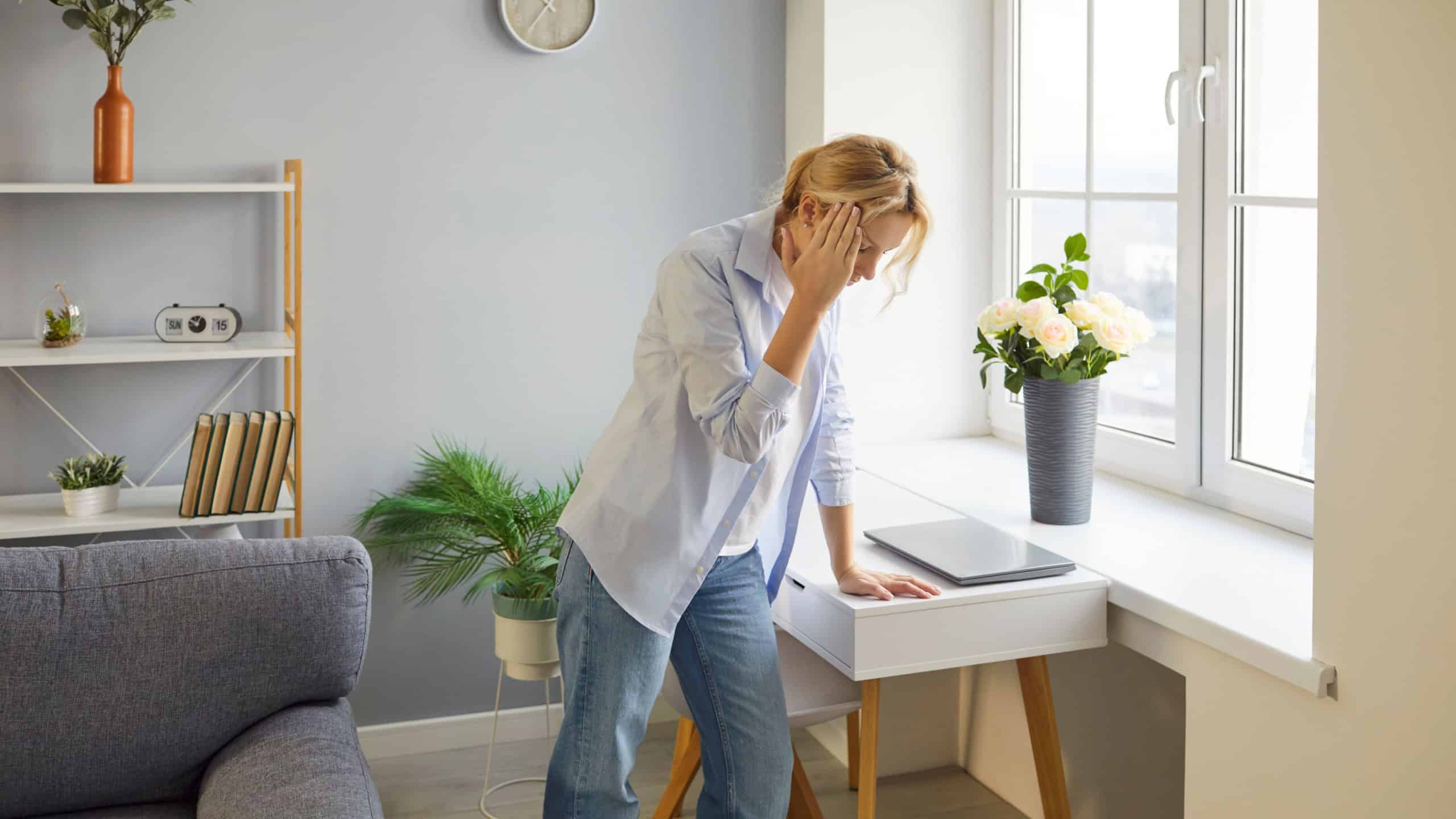 Weary blonde woman standing by window, resting hand on forehead, feeling unwell, suffering from dizziness. Weak girl experiencing vertigo, exhaustion or heat discomfort, struggling to stay conscious.