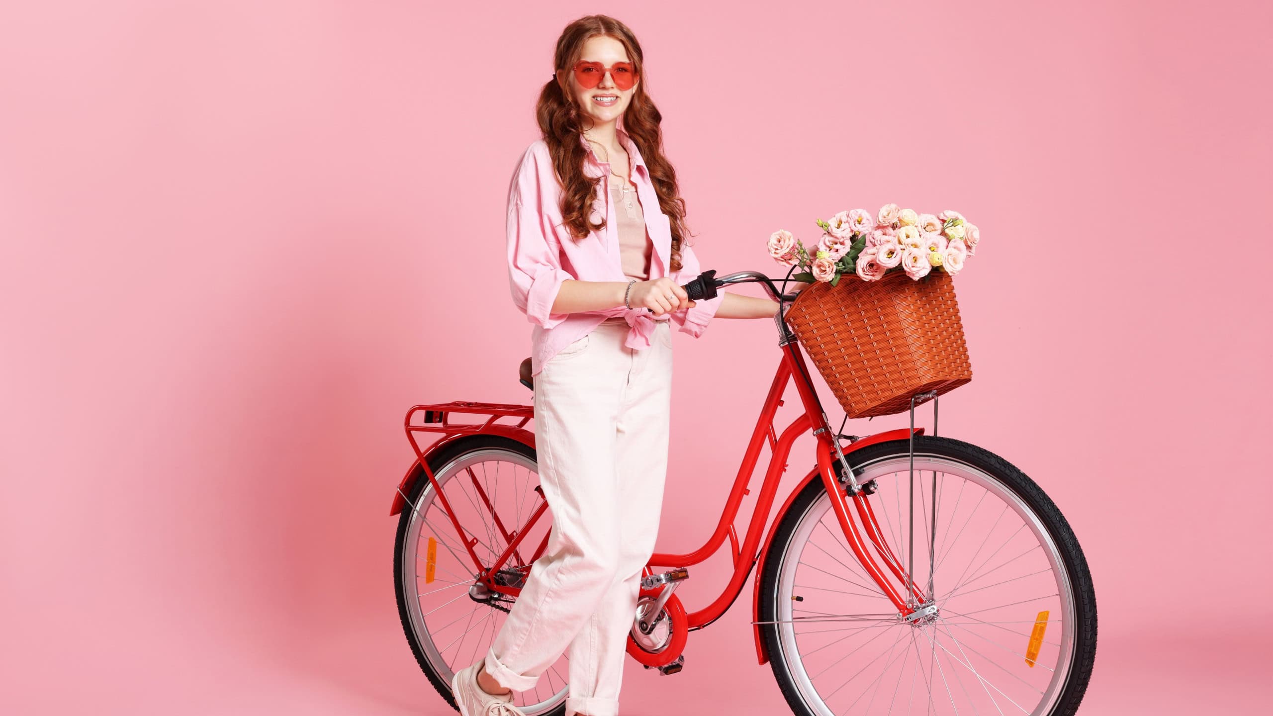 Smiling teenage girl in sunglasses and bicycle with basket of flowers against pink background. Space for text