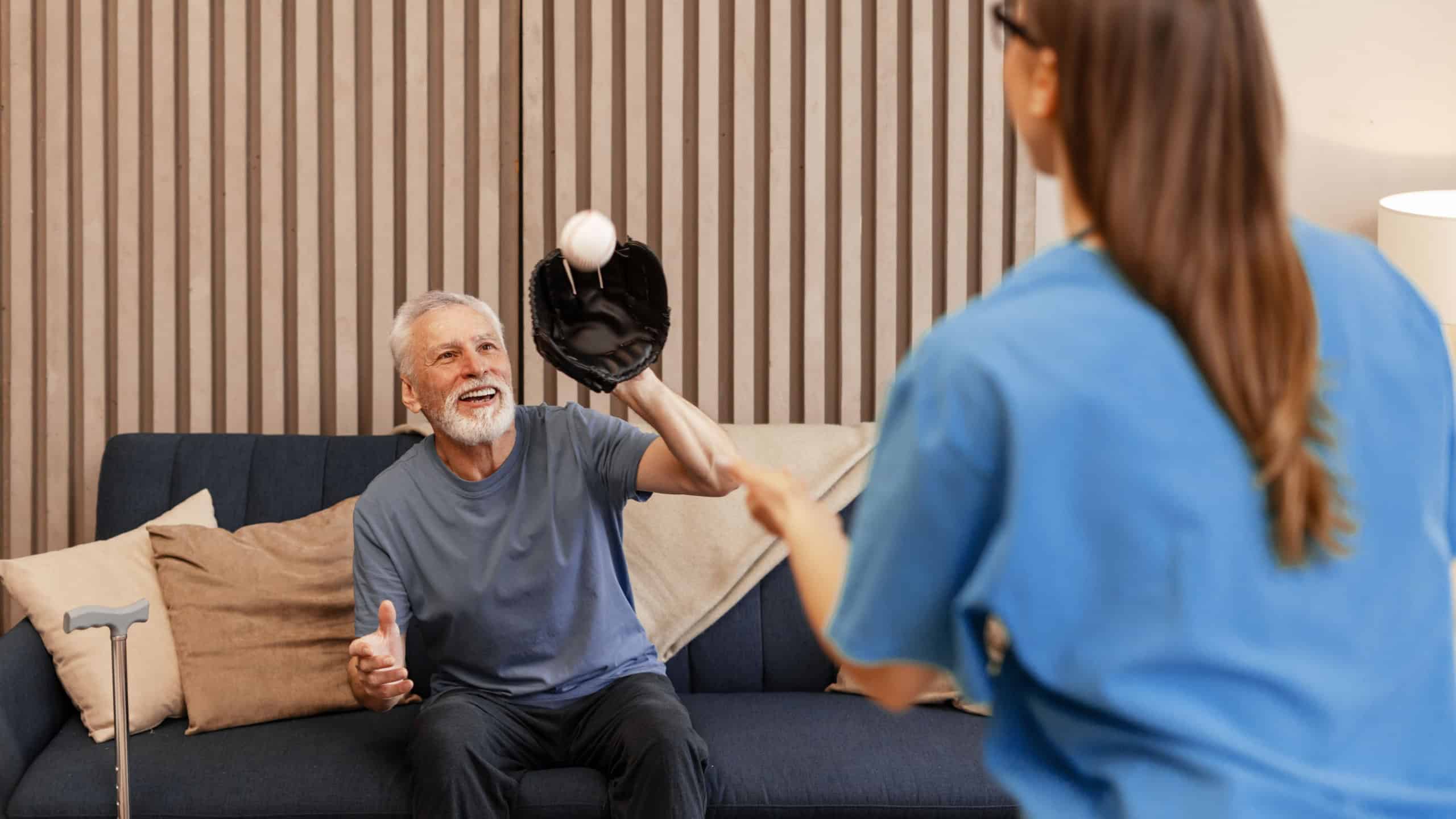 Senior man with walking cane is catching a baseball with his nurse, playing a game in the living room of a modern home, improving his health and well-being. Rehabilitation concept
