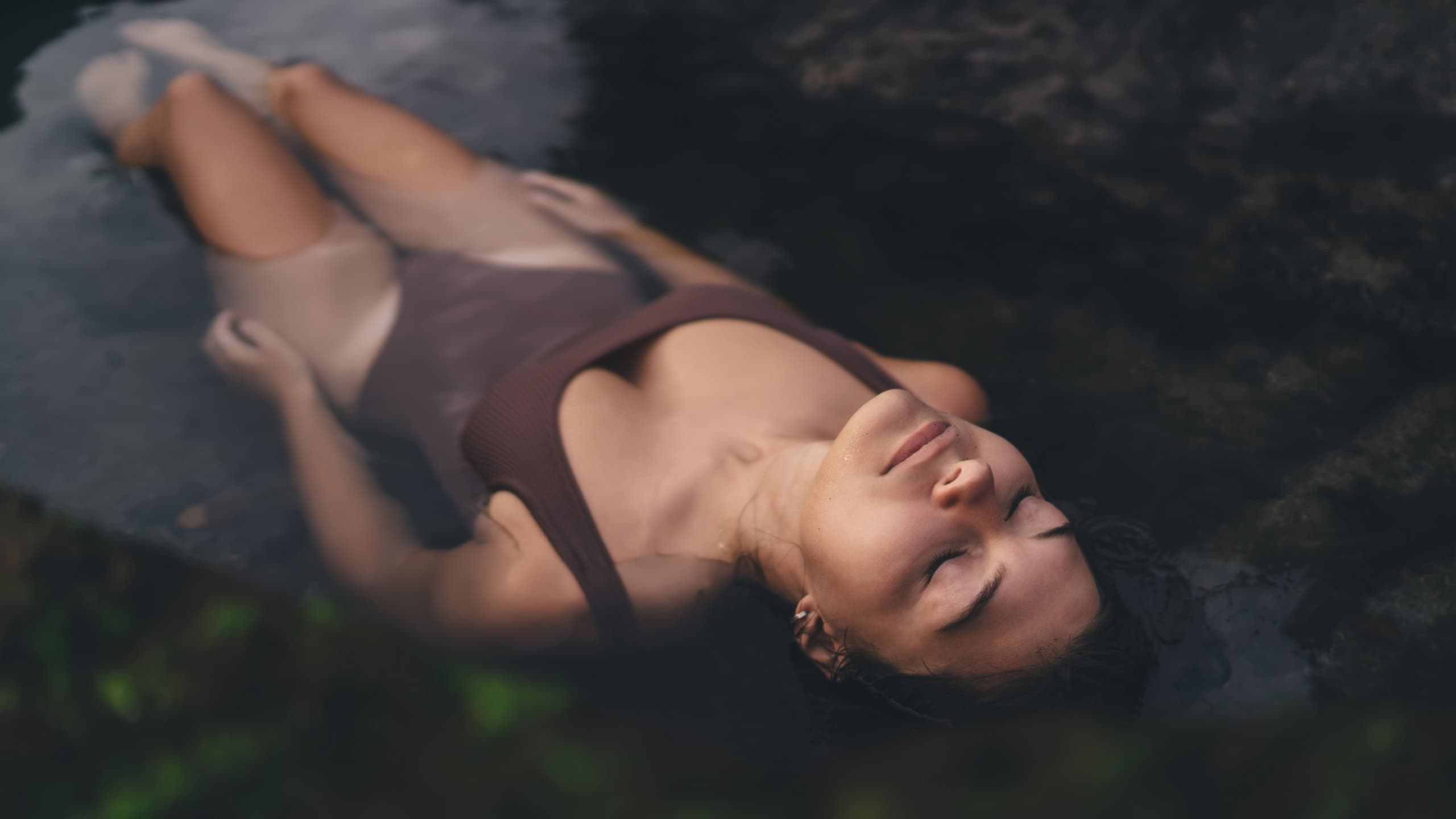 Woman floats on back with relaxed expression, body merging into rock pool, symbolizing emotional surrender and contrast to artificial, filtered perfection of virtual life.