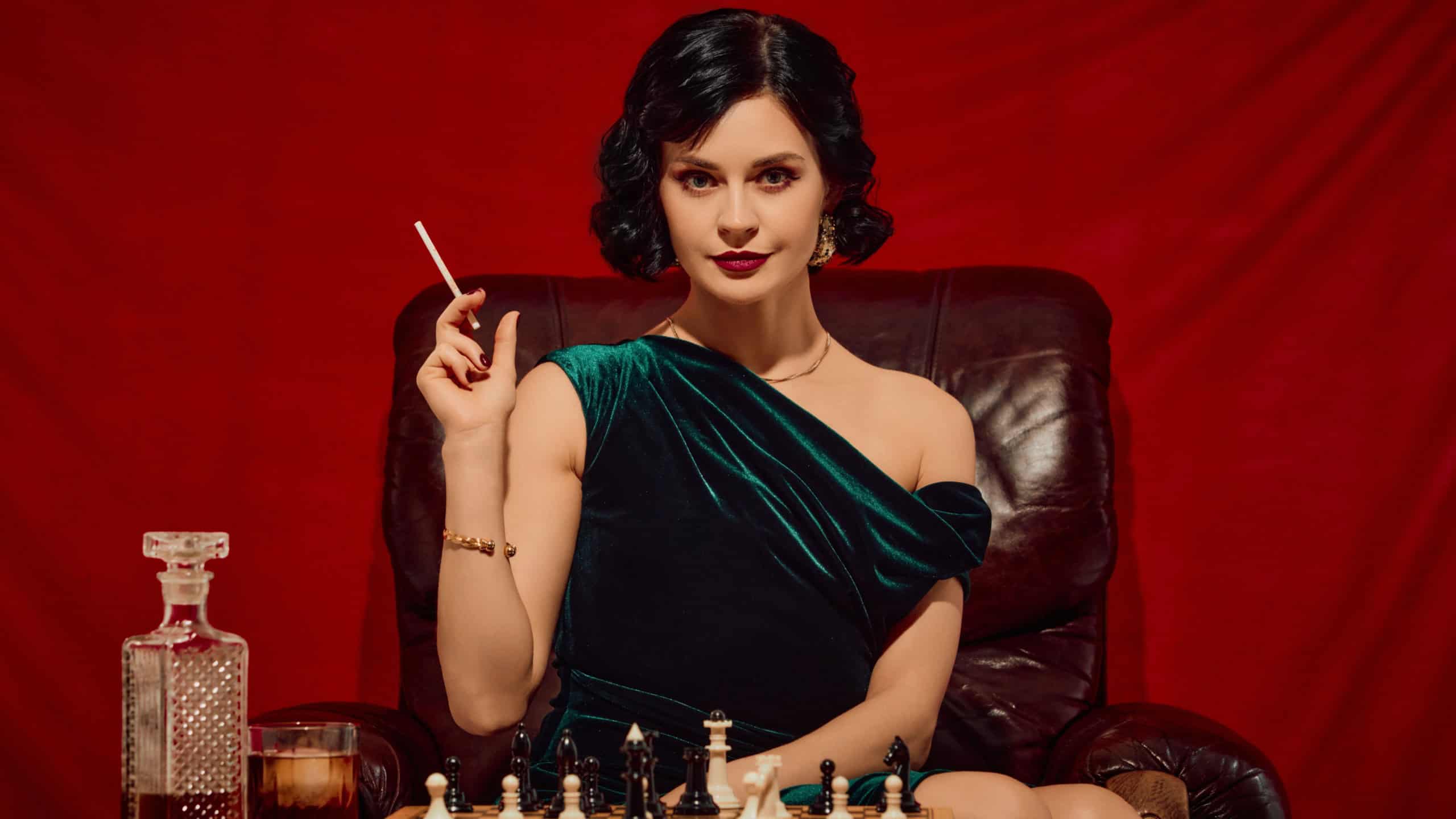 Woman in velvet dress holding cigarette and glass sitting at chessboard with intense expression. Concept of power pose, luxury retro branding, strategy visuals, elegant female lifestyle.