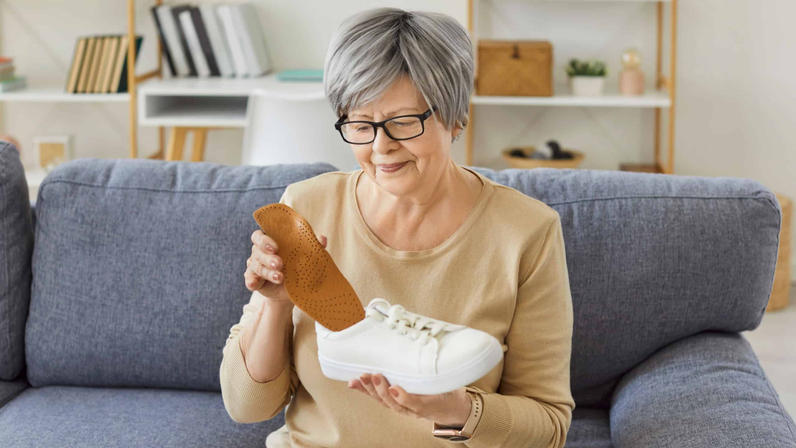 Smiling elderly woman sitting on couch and holding white sneaker with orthopedic insole at home. Female pensioner using foot care accessories for pain relief, comfort and daily walking support.