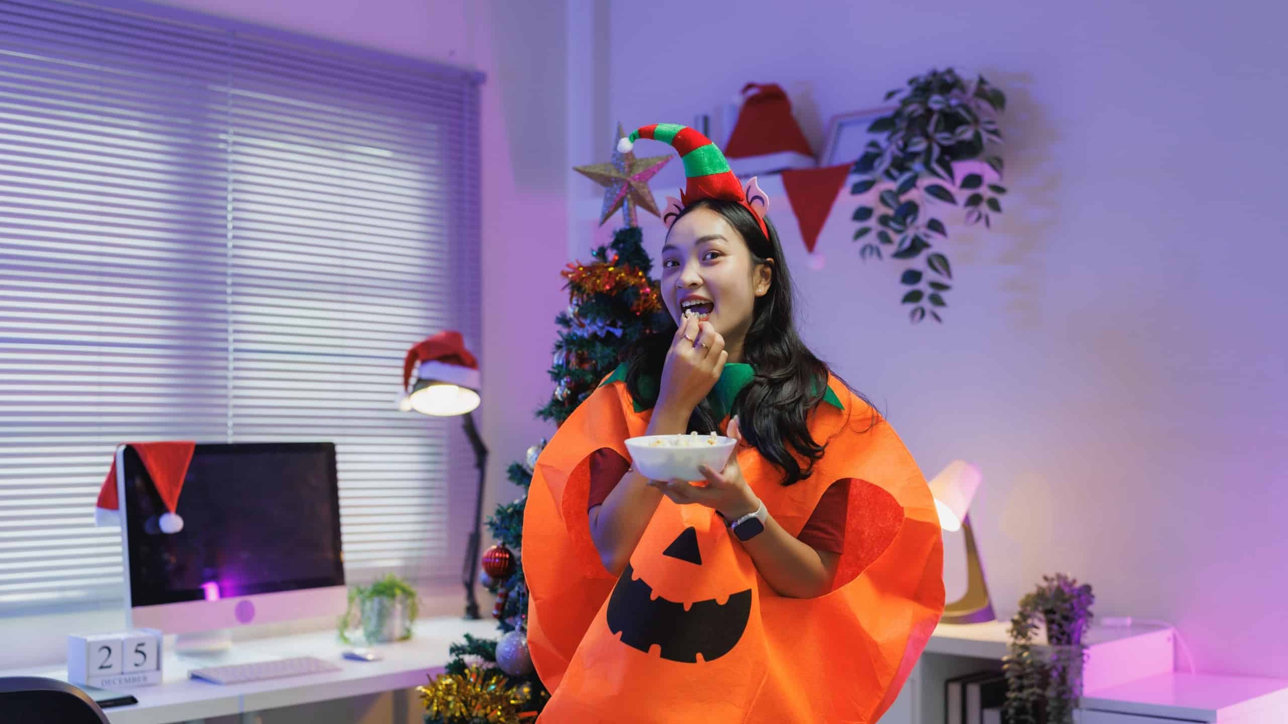 Young woman in a pumpkin costume and elf hat eating popcorn, celebrating christmas and halloween in a festive home office