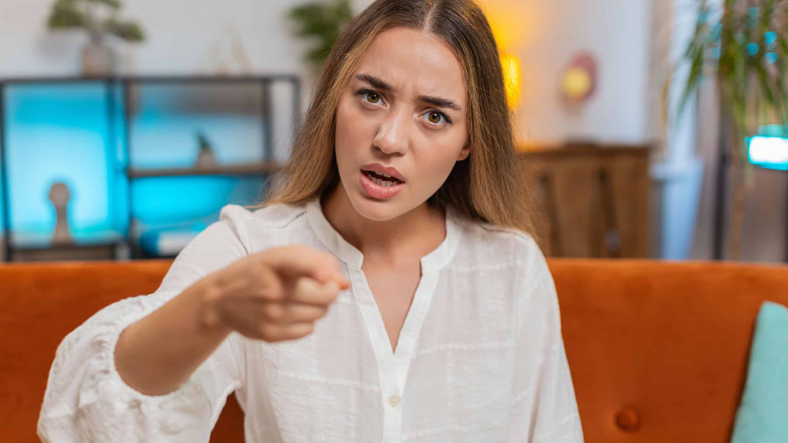 Furious young Caucasian woman sitting on sofa couch at home, angrily pointing at you through the camera, displeased and fuming. Adult Girl in serious blame mode, upset, and ready to explode. Lifestyle