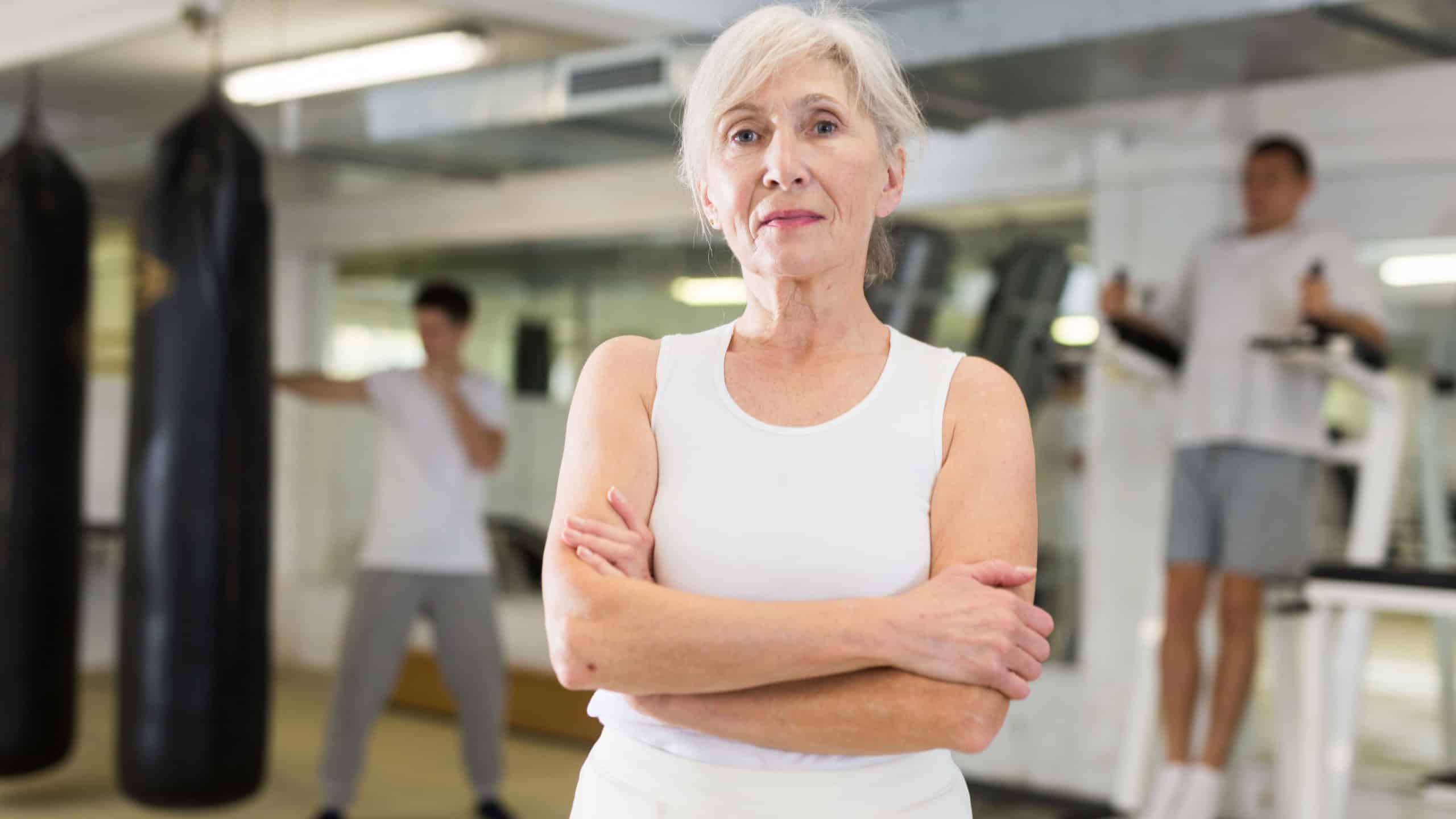 Waist-up portrait of senior woman standing in gym with crossed hands.