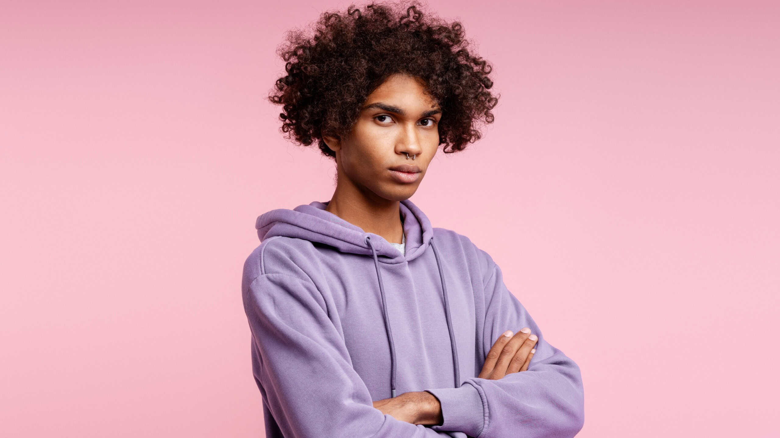 Young black man with curly hair and a nose ring standing confidently with arms crossed on a pink background