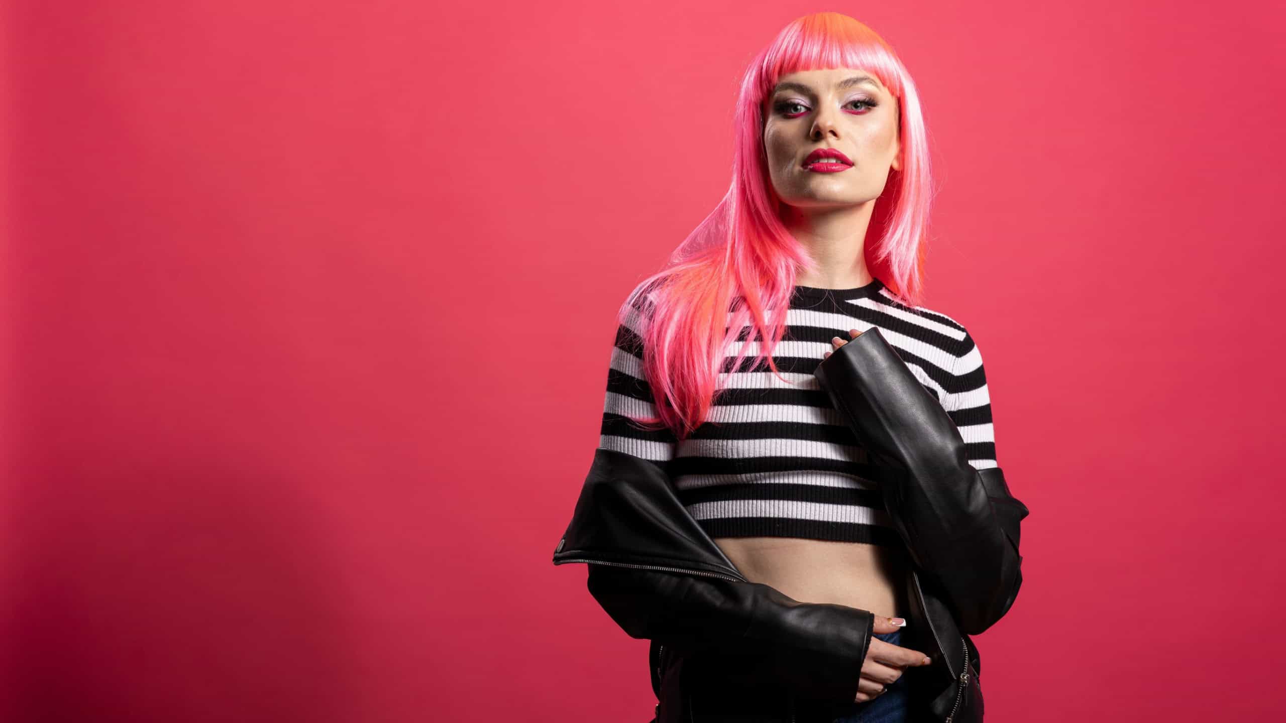 Confident fashion model poses freely, blending elegance with punk flair to express modern rebellious beauty. Young woman with bold pink hair rocks edgy leather jacket look in stylish studio portrait.