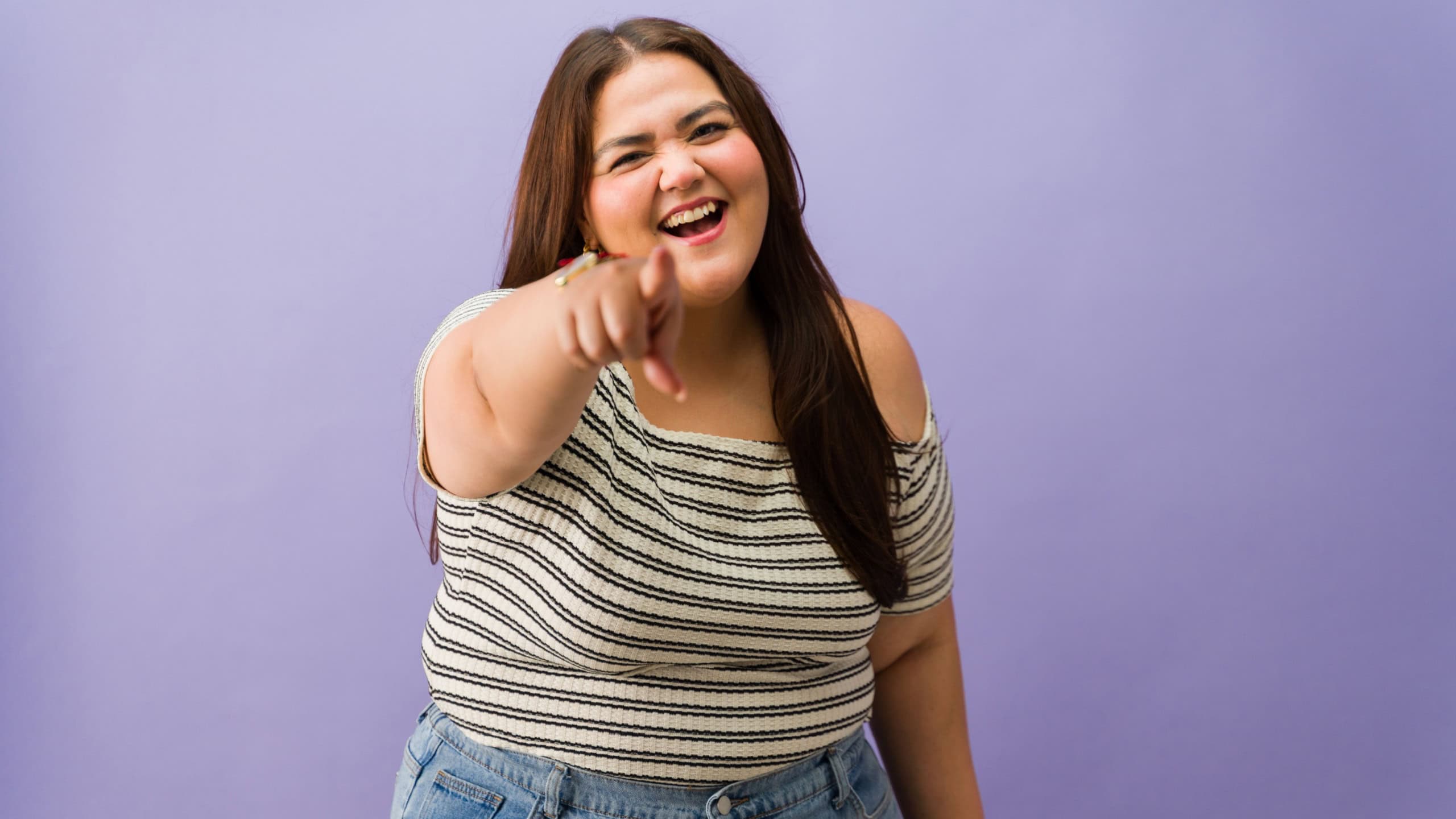 Cute plus size woman smiling, laughing and playfully pointing a finger directly at the viewer in a studio