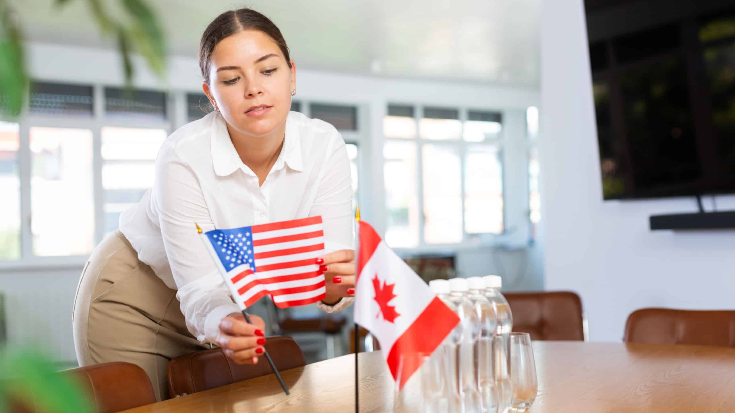 Young woman in business clothes puts flags of USA and Canada on negotiating table in office..