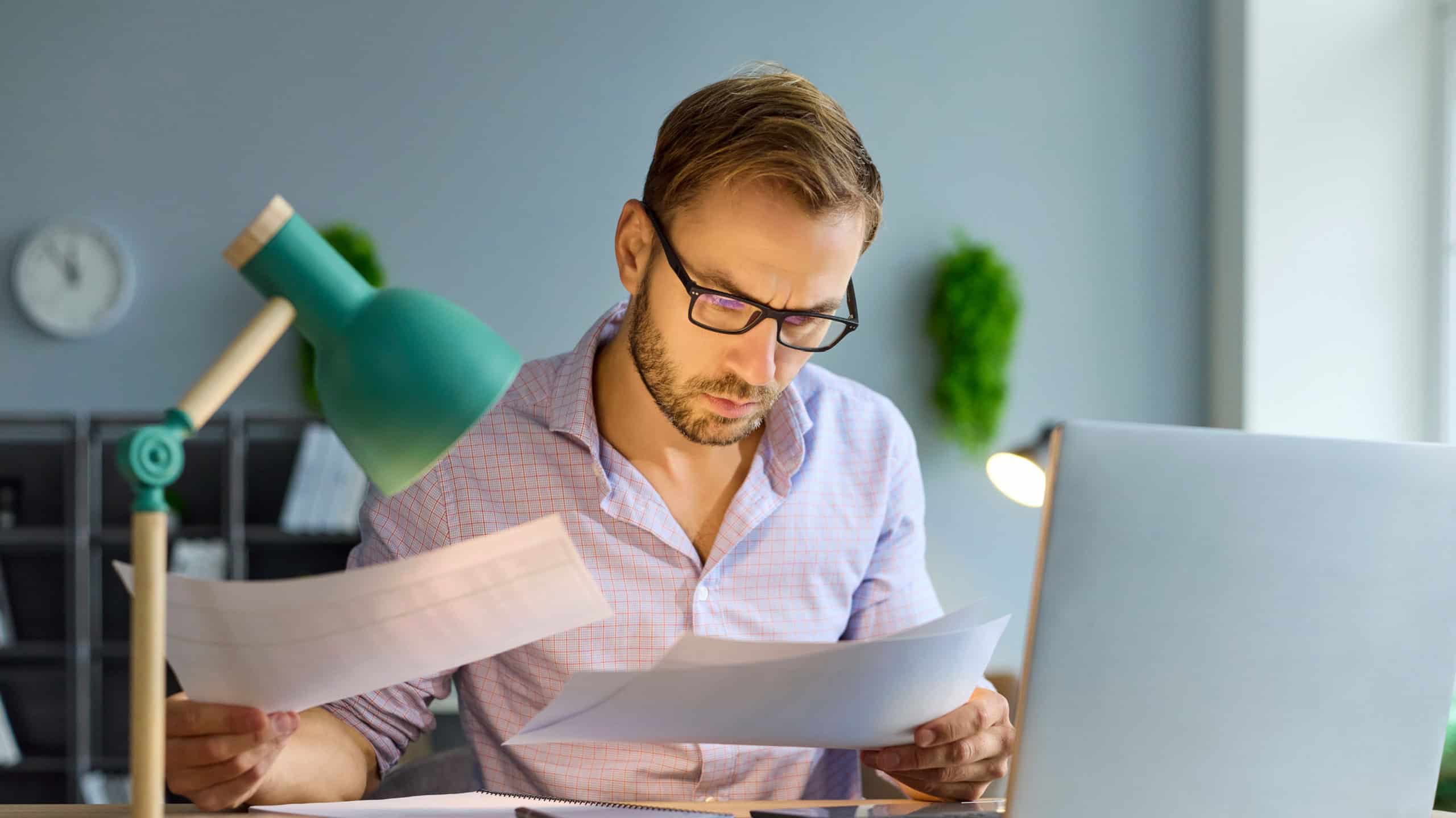 Busy accountant looking at business documents, financial reports to review paperwork at office. Careful audit with finance analysis of workaholic man in glasses using laptop, sitting at desk with lamp