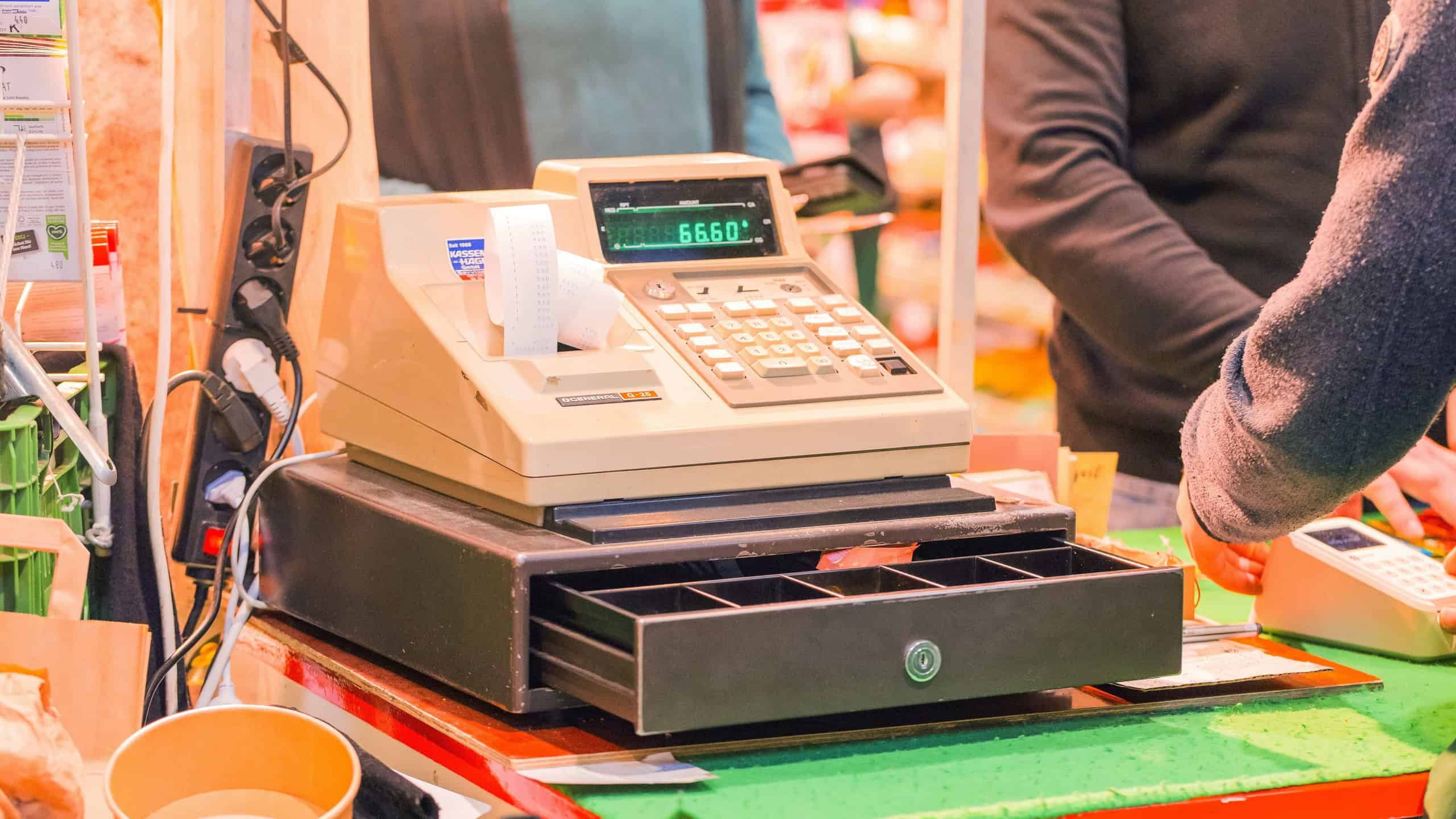Cash register displayed on a market stall, featuring an open drawer and various colorful items in the background, creating a lively atmosphere.