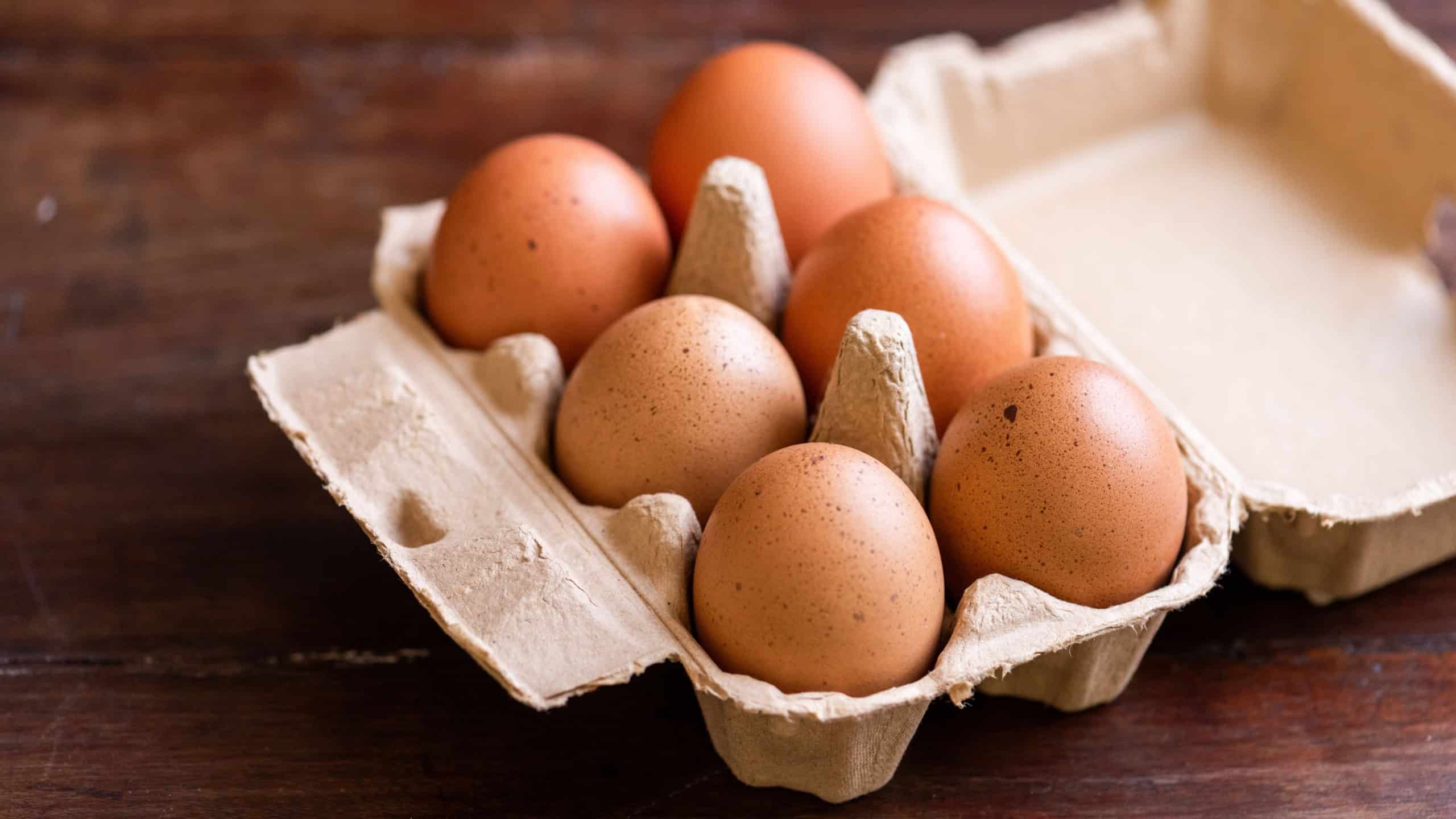 A carton of six brown eggs on a wooden table. The eggs are neatly arranged in a cardboard egg carton, showcasing their natural speckled shells.