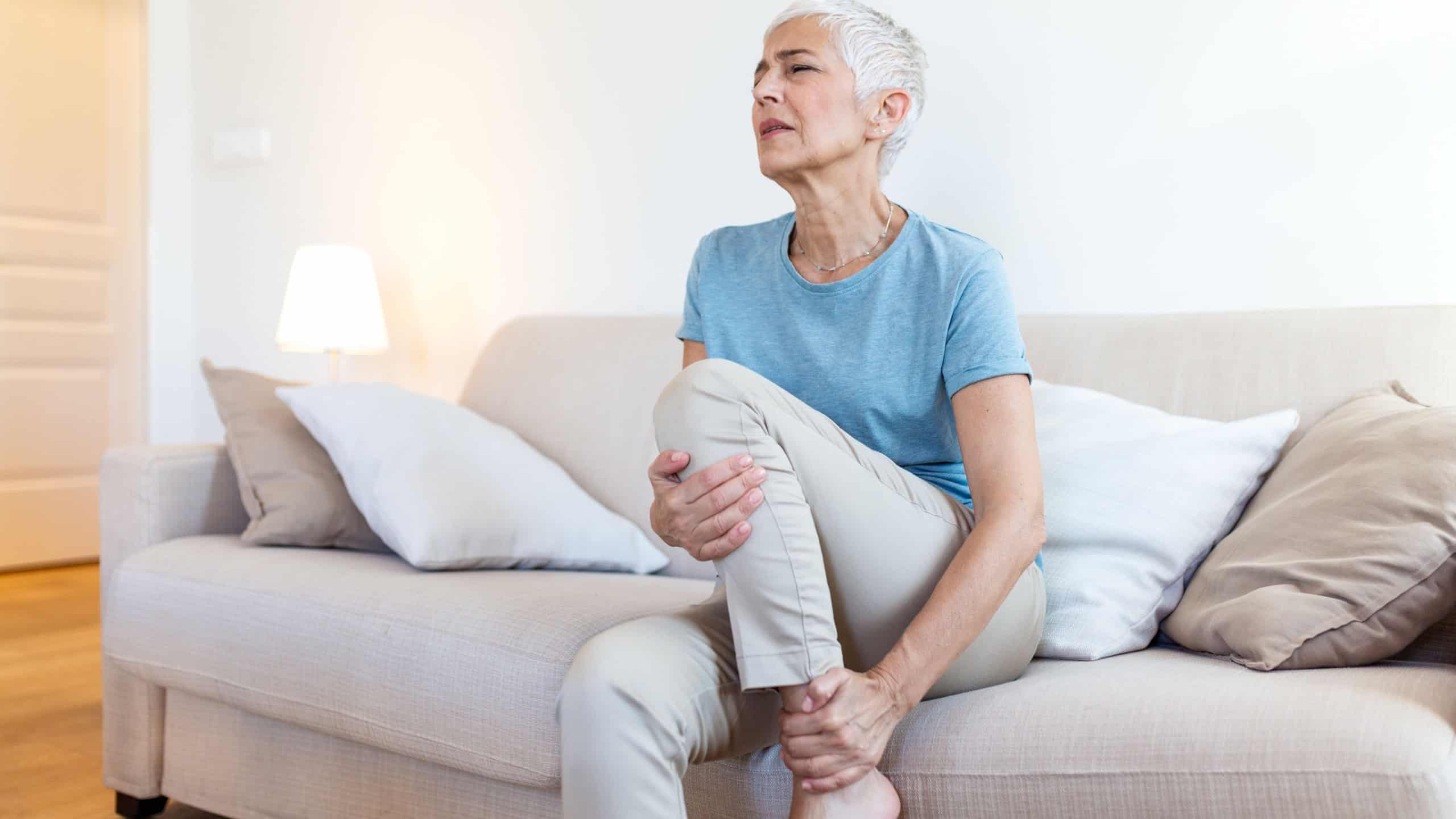 Knee pain in senior woman. Mature lady sitting on sofa and holding painful leg with hands. Healthcare and medicine concept. Elderly person suffering from joint ache, arthritis, or physical injury.