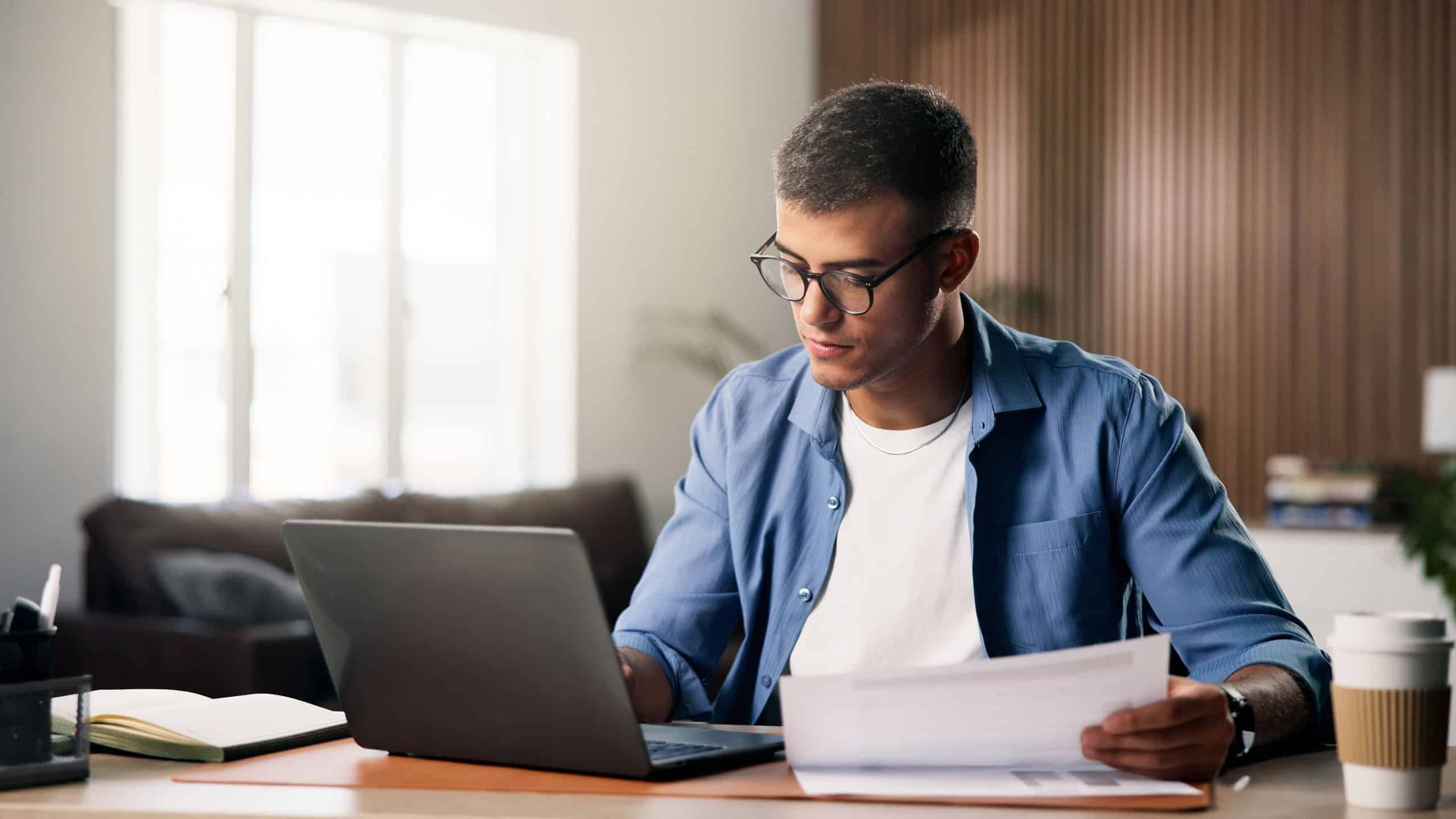 Documents, laptop and remote work with man in home study for planning, research or review. Computer, paperwork and report with freelance employee at desk in apartment for small business startup