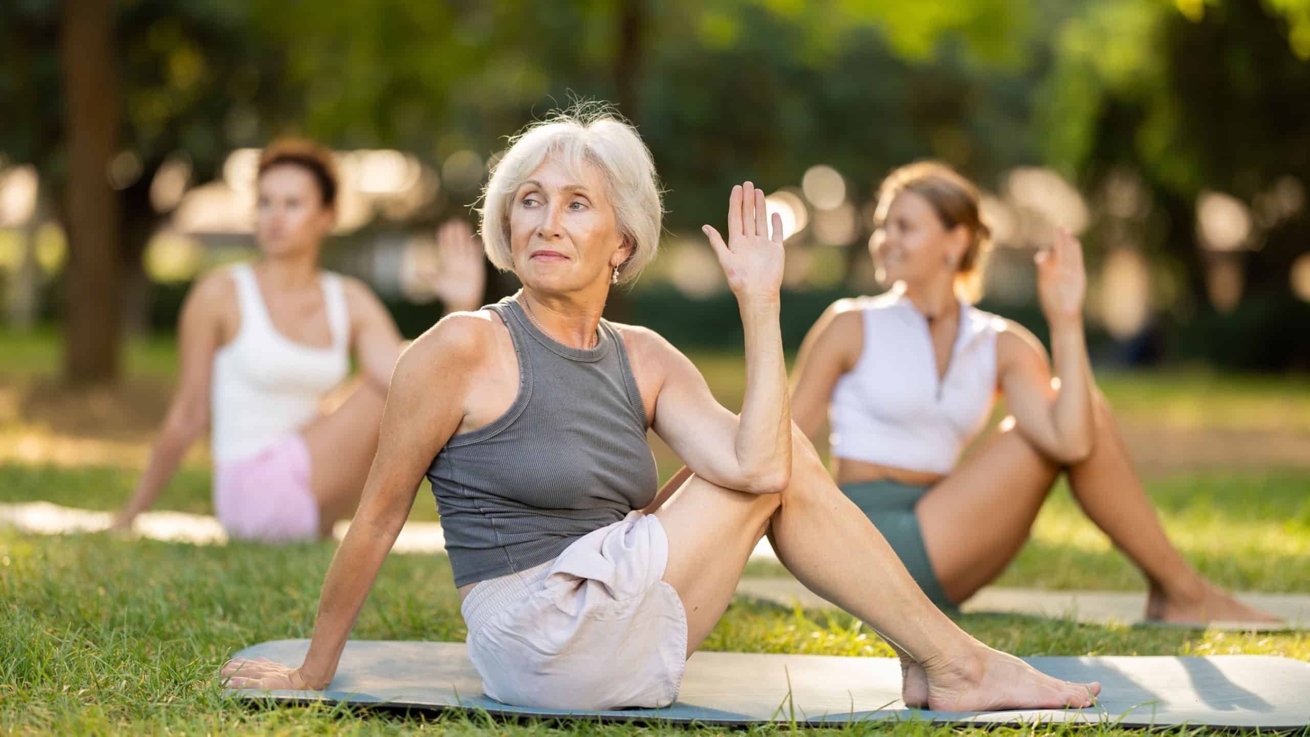 Sedulous old lady and group of women doing spinal twist pose of yoga on mat in modern light city park together