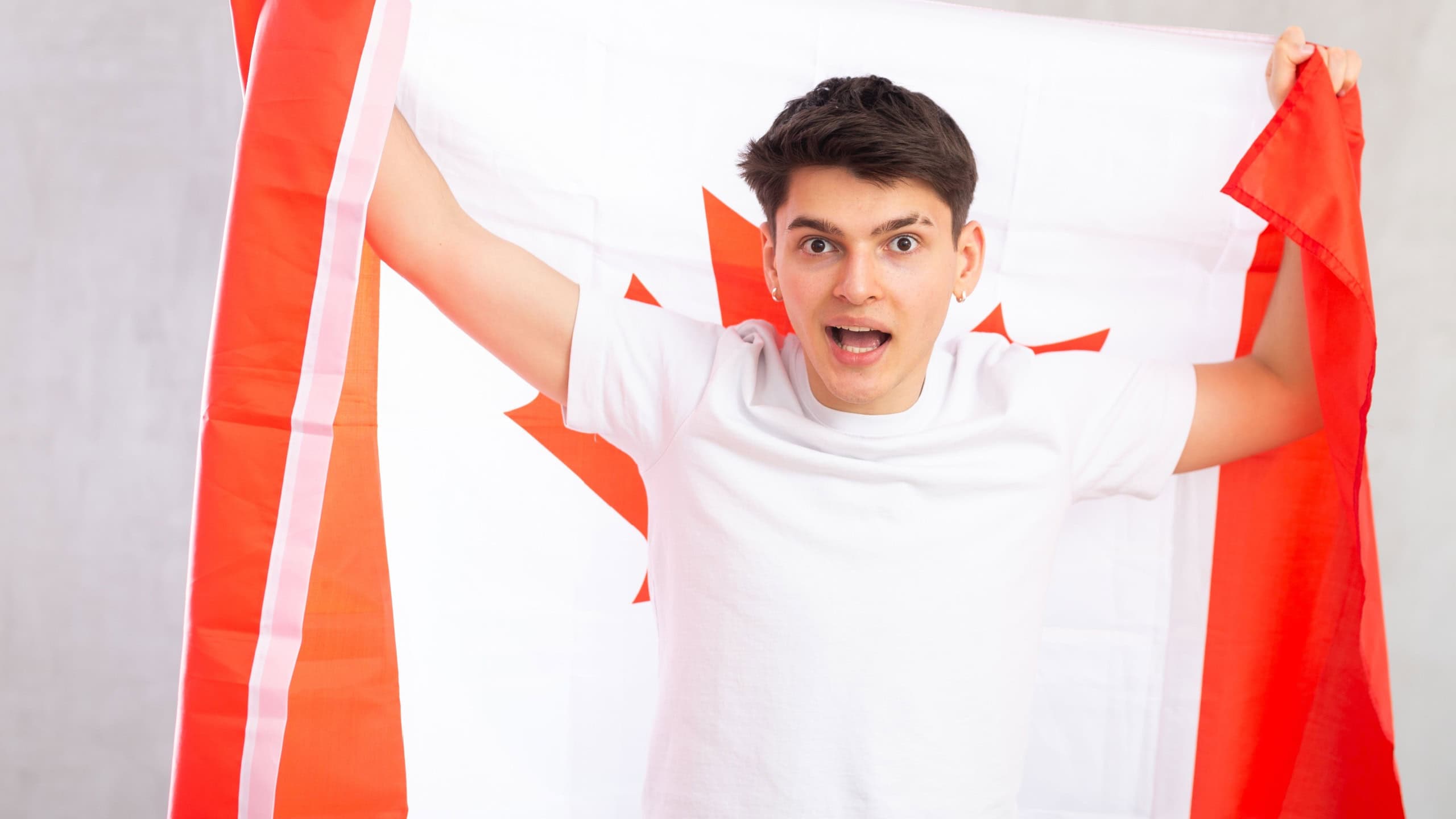 Young surprised guy teenager posing with big flag of Canada in his hands in studio