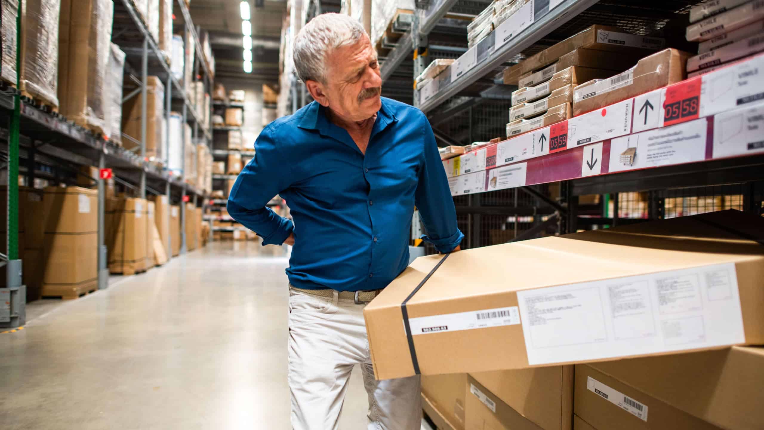 Older man in a blue shirt and white pants experiencing back pain while lifting a large box in a warehouse aisle filled with shelves and packages.