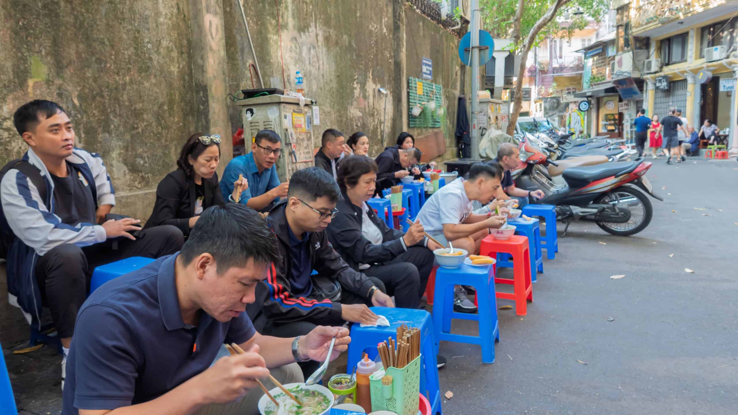 Hanoi, Vietnam - Nov 20, 2024: Busy sidewalk scene with many people eating pho on small stools along a street in Hanoi Old Quarter with parked motorbikes.