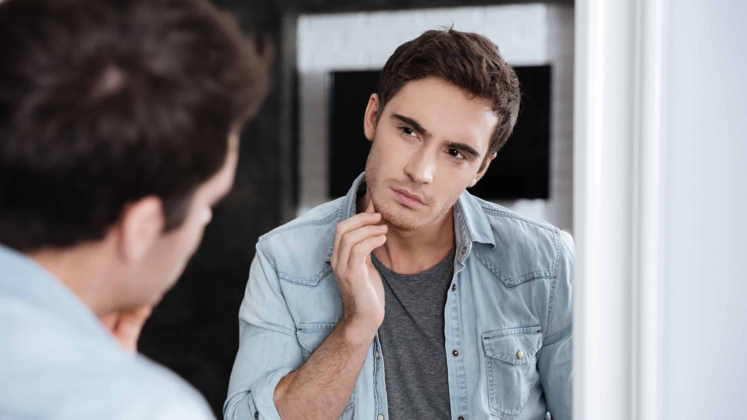 Young man touching his face with hand and gazing confidently while standing in front of the mirror