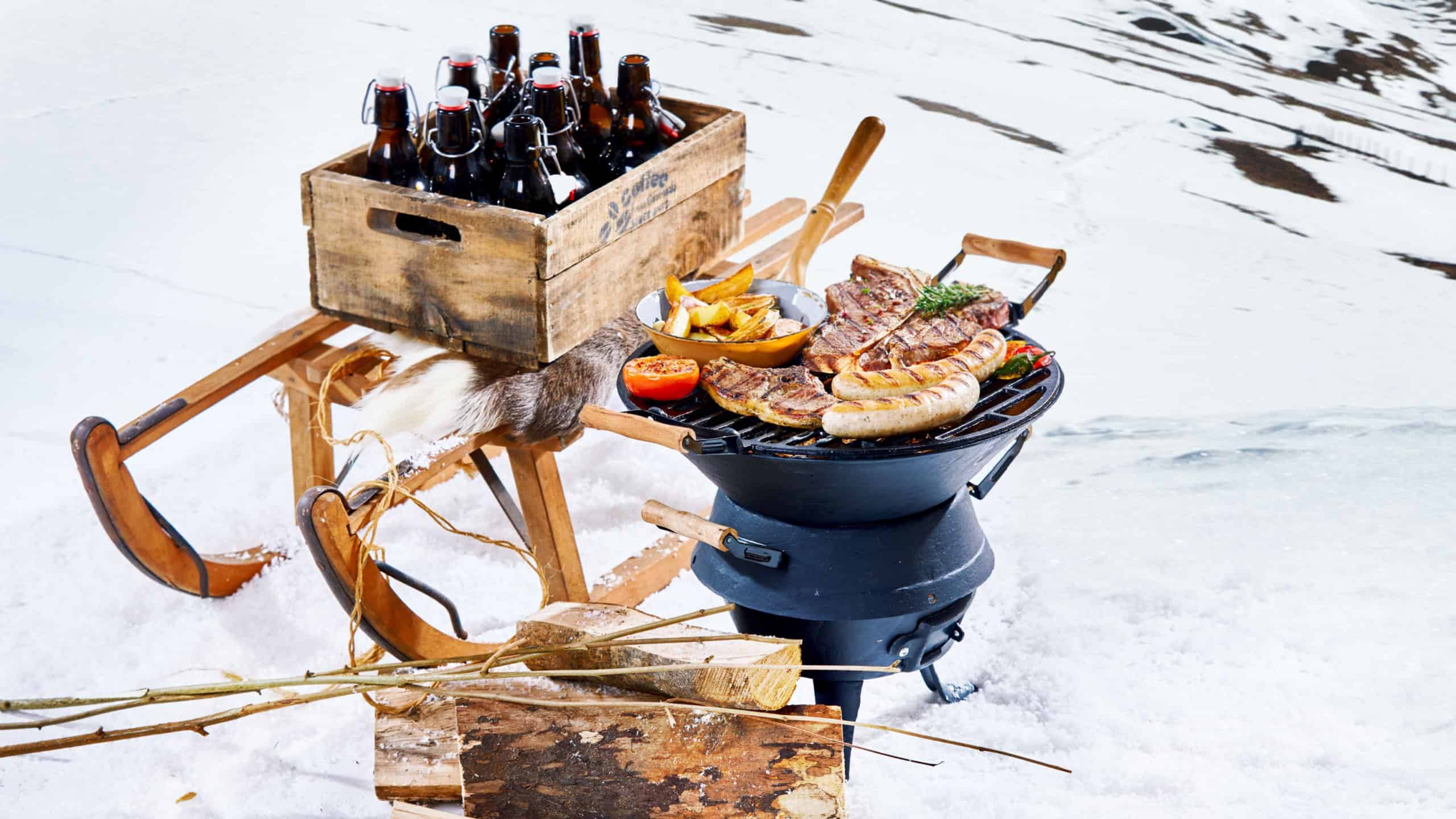 Snowy winter barbecue outdoors in the cold with a crate of beer bottles on a sled and spicy sausages and T-bone steaks grilling on a portable BBQ