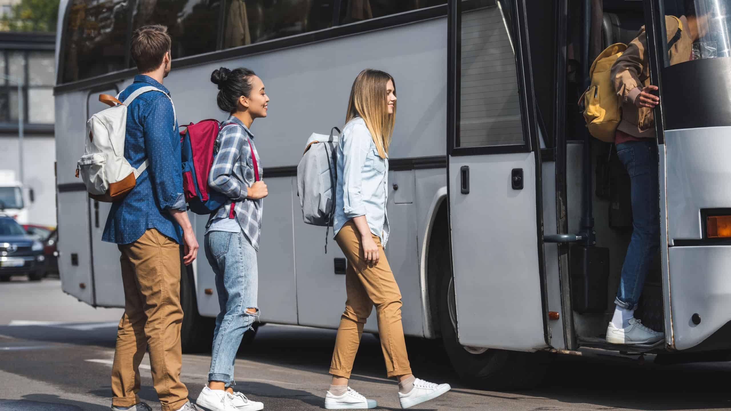 young smiling tourists with rucksacks walking into travel bus at city street 