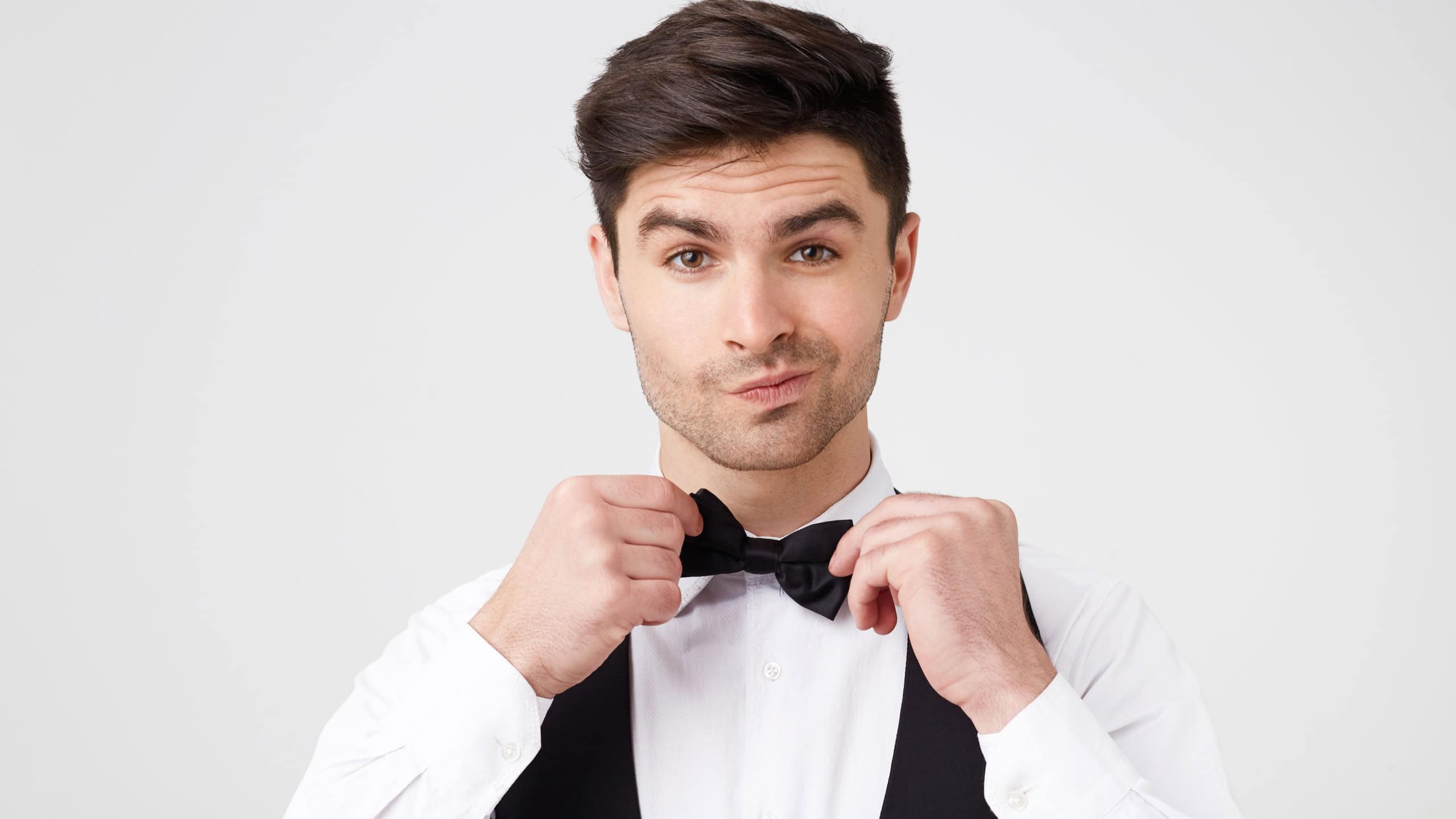 Self-confident brunet dressed in a smart suit straightens black butterfly has no doubt that he looks great and the date will go superbly, isolated white studio background