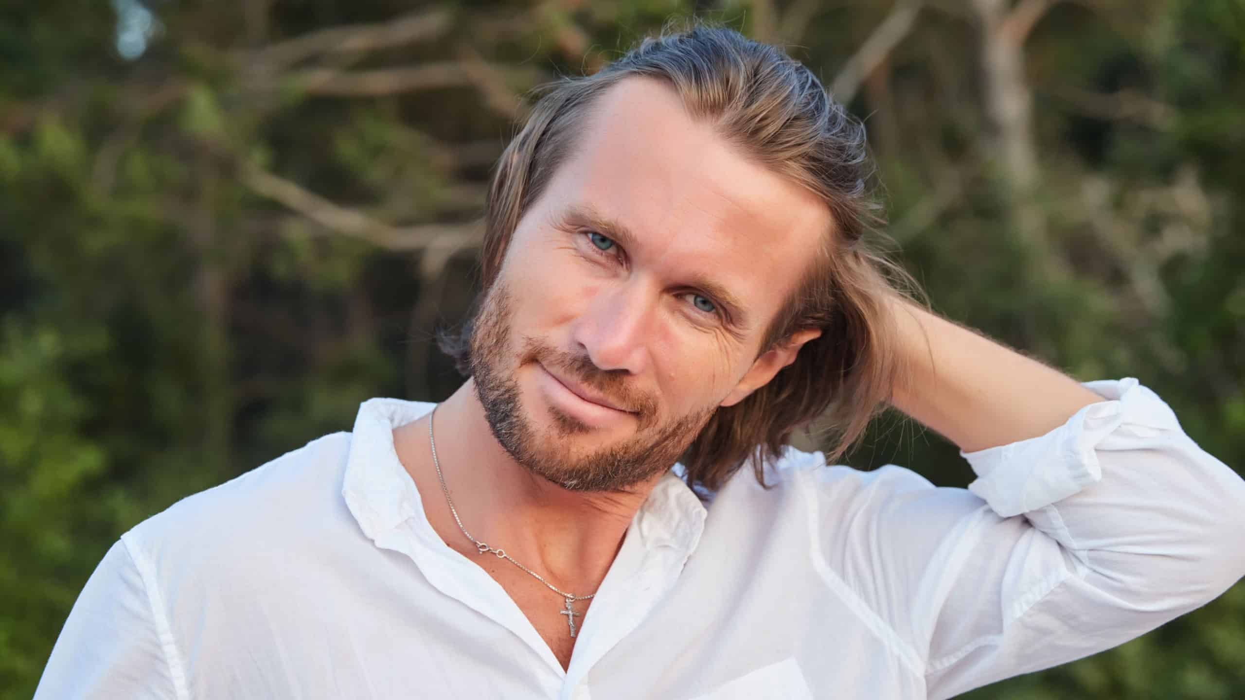 Portrait of the young handsome brutal man with long hair, the blue eyes, an easy bristle, he is dressed in a white shirt with a short sleeve, green trees on a background, light of a sunset