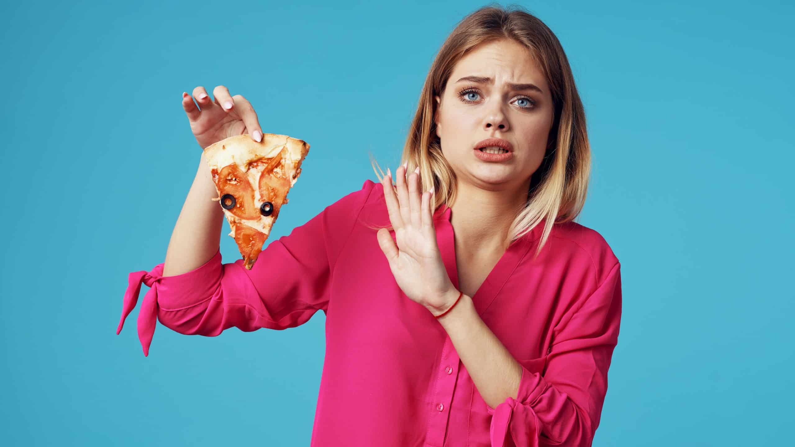A woman in a pink shirt does not want to eat fresh pizza in her hand on a blue background