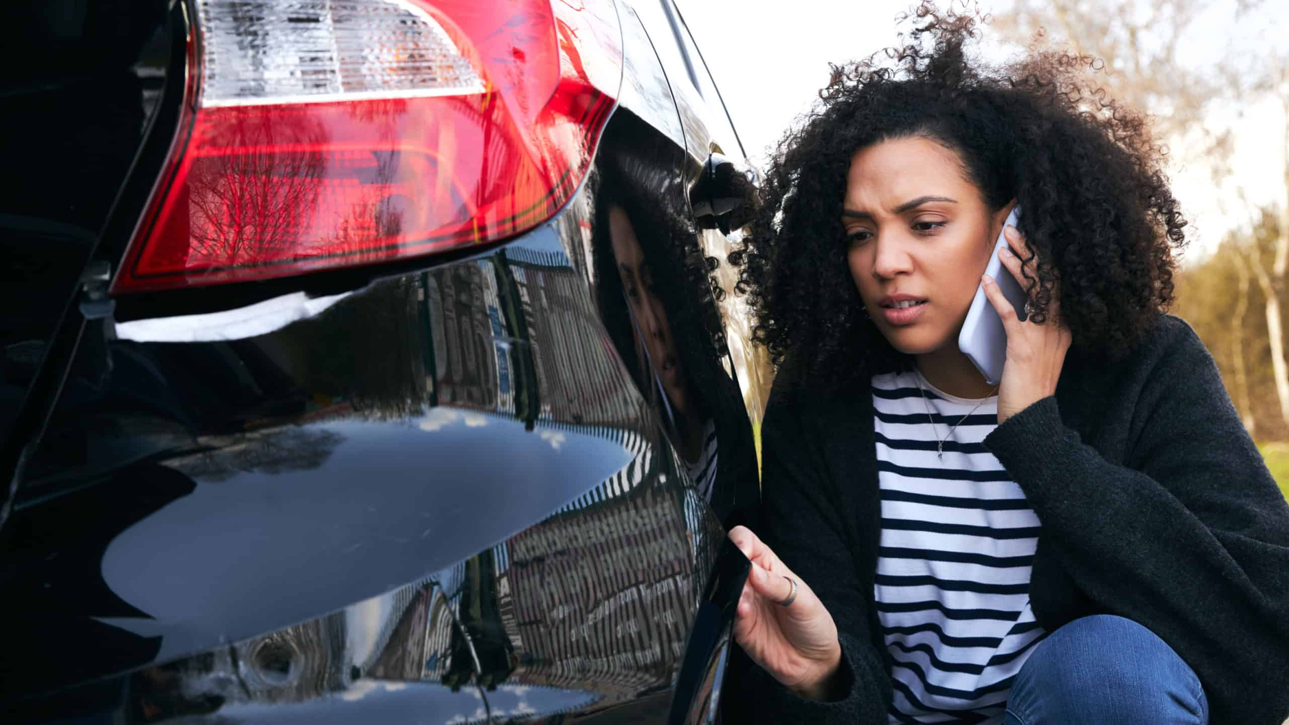Angry young woman standing by vandalised car in car park reporting damage to insurance company using mobile phone