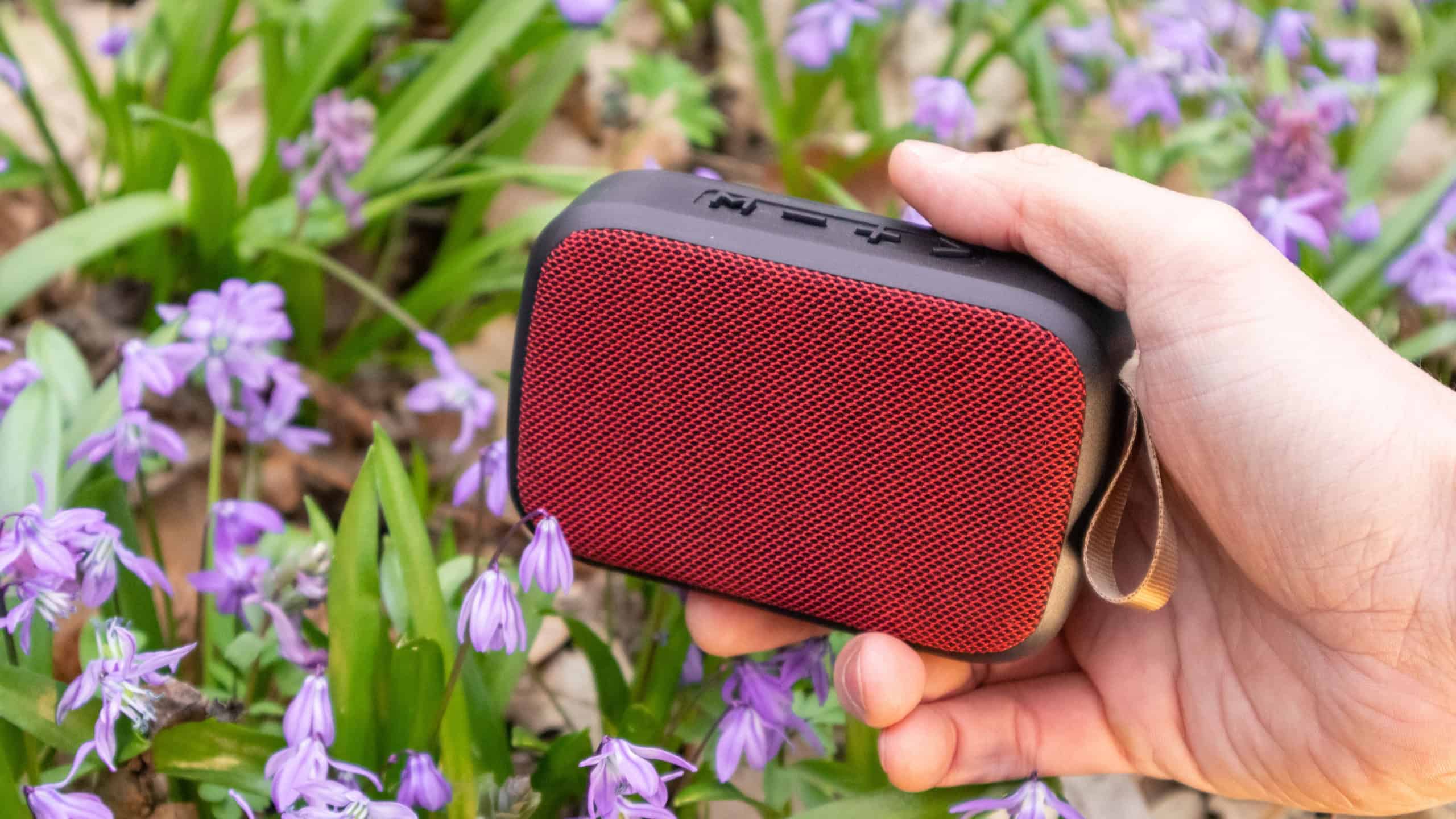 Hand holding red portable audio speaker in wild nature close-up. Blooming wild spring lawn purple flowers background. Take music with you outdoors