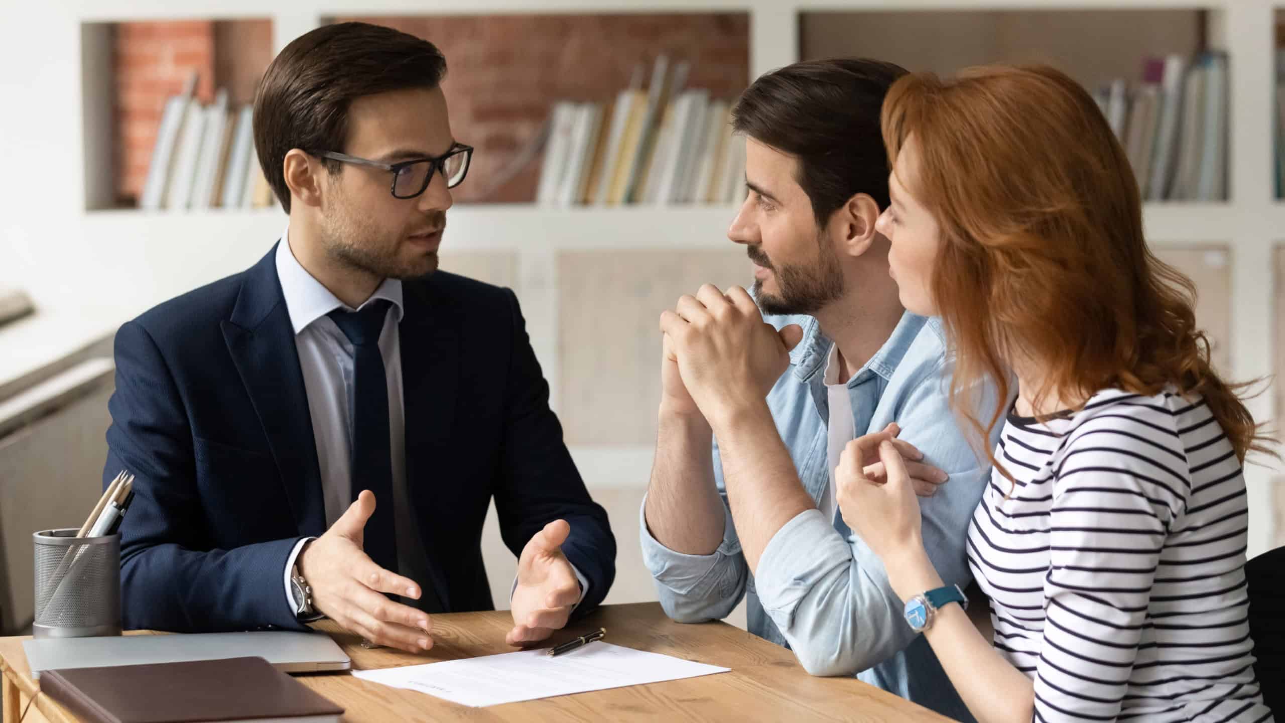Professional male lawyer financial advisor consulting happy family couple clients in modern office. Interested young customers meeting realtor broker bank worker, discussing agreement or deal.