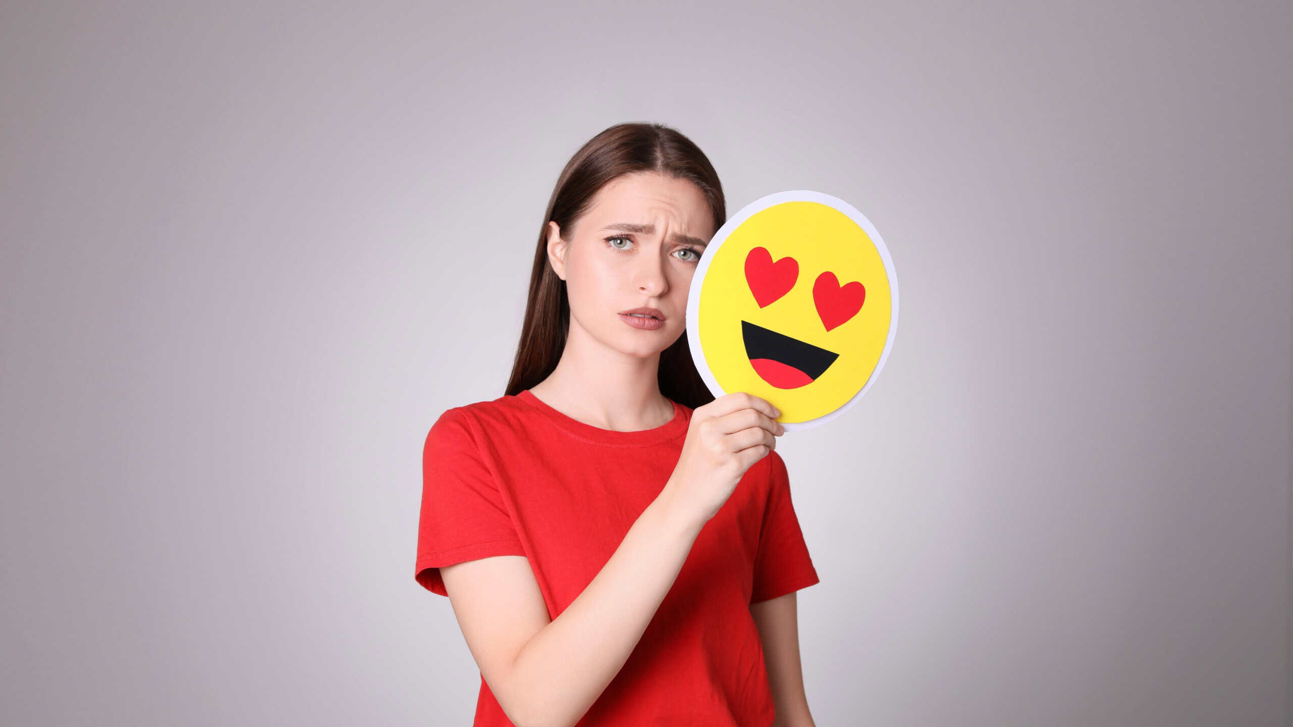 Sad young woman covering face with heart eyes emoji on grey background
