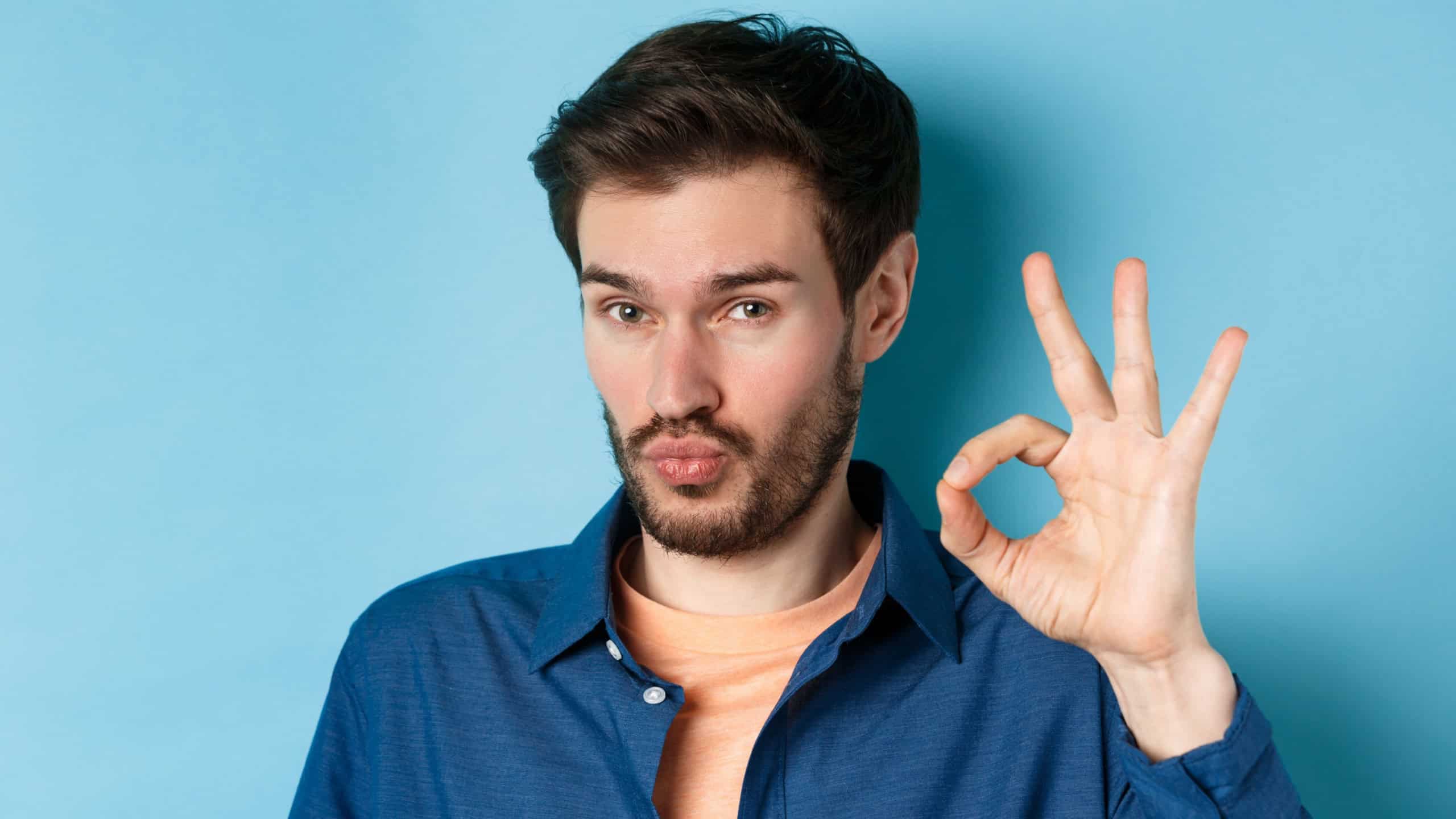 Image of silly young man pucker lips and show okay gesture, praise good thing, compliment excellent choice, standing on blue background