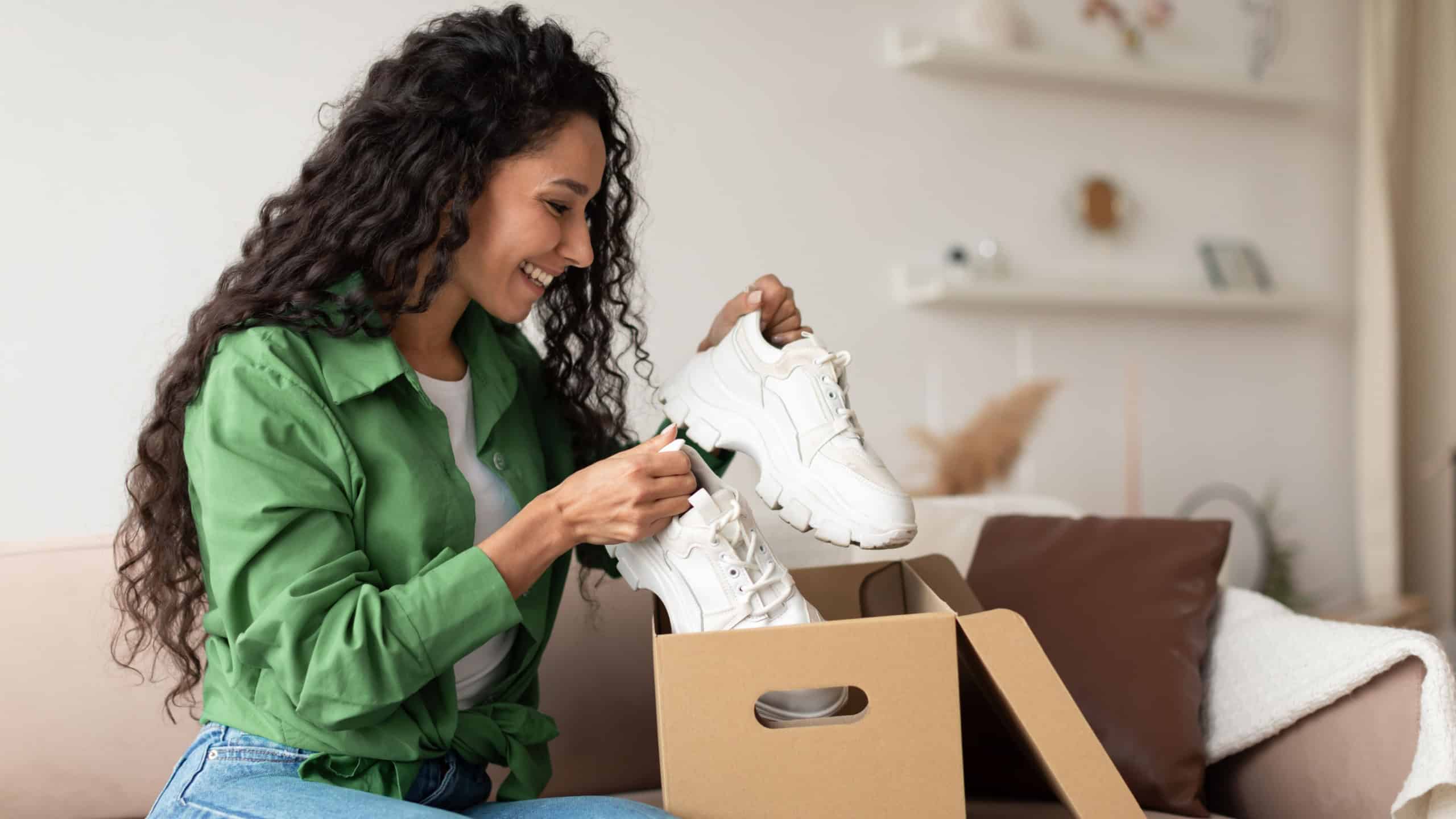 Delivery Concept. Happy Female Buyer Holding Footwear Unpacking Cardboard Box Sitting On Couch At Home. Joyful Customer Woman Receiving Shoes After Successful Online Shopping