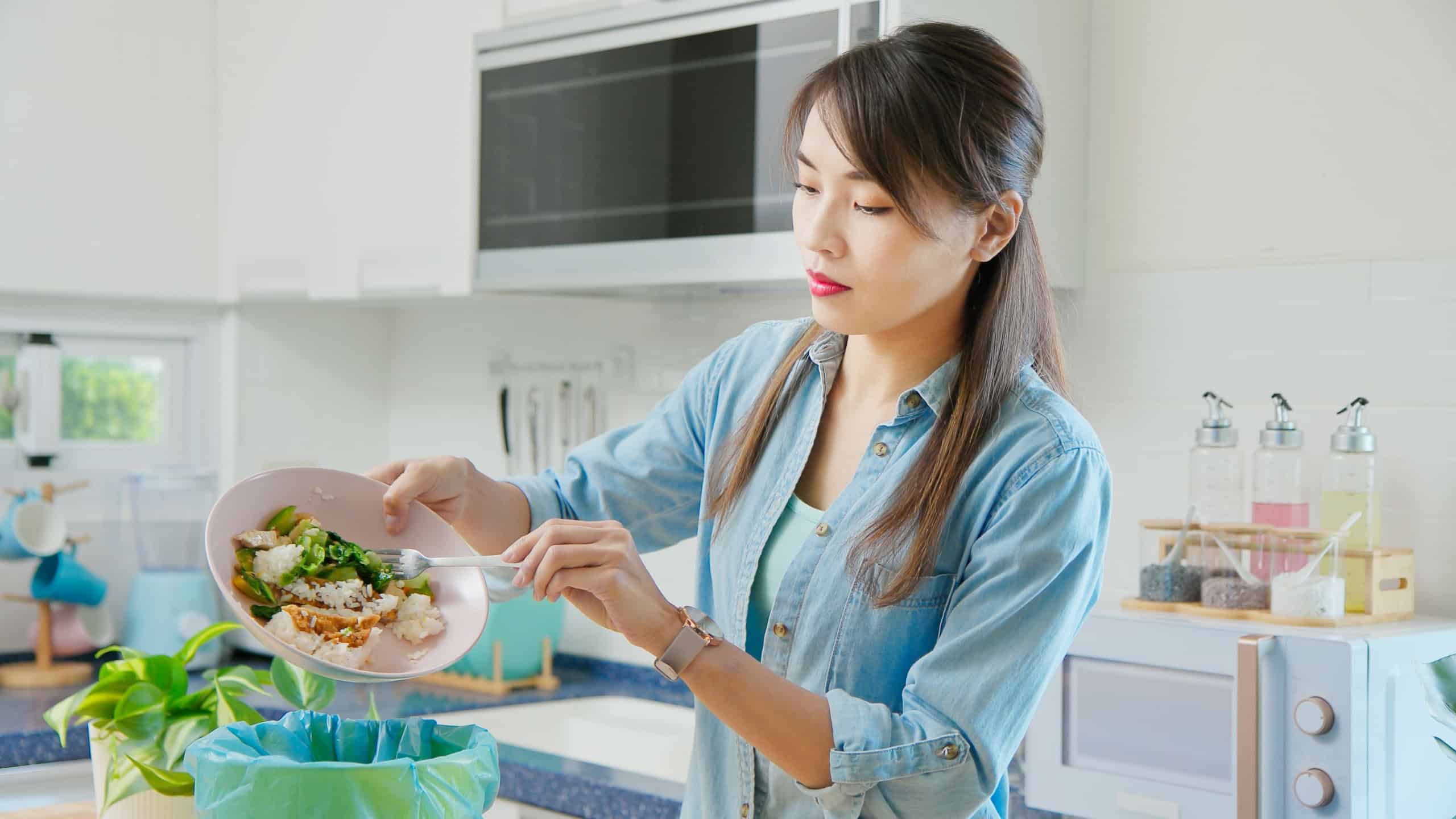 asian woman scraping food leftovers or waste into kitchen bucket at home