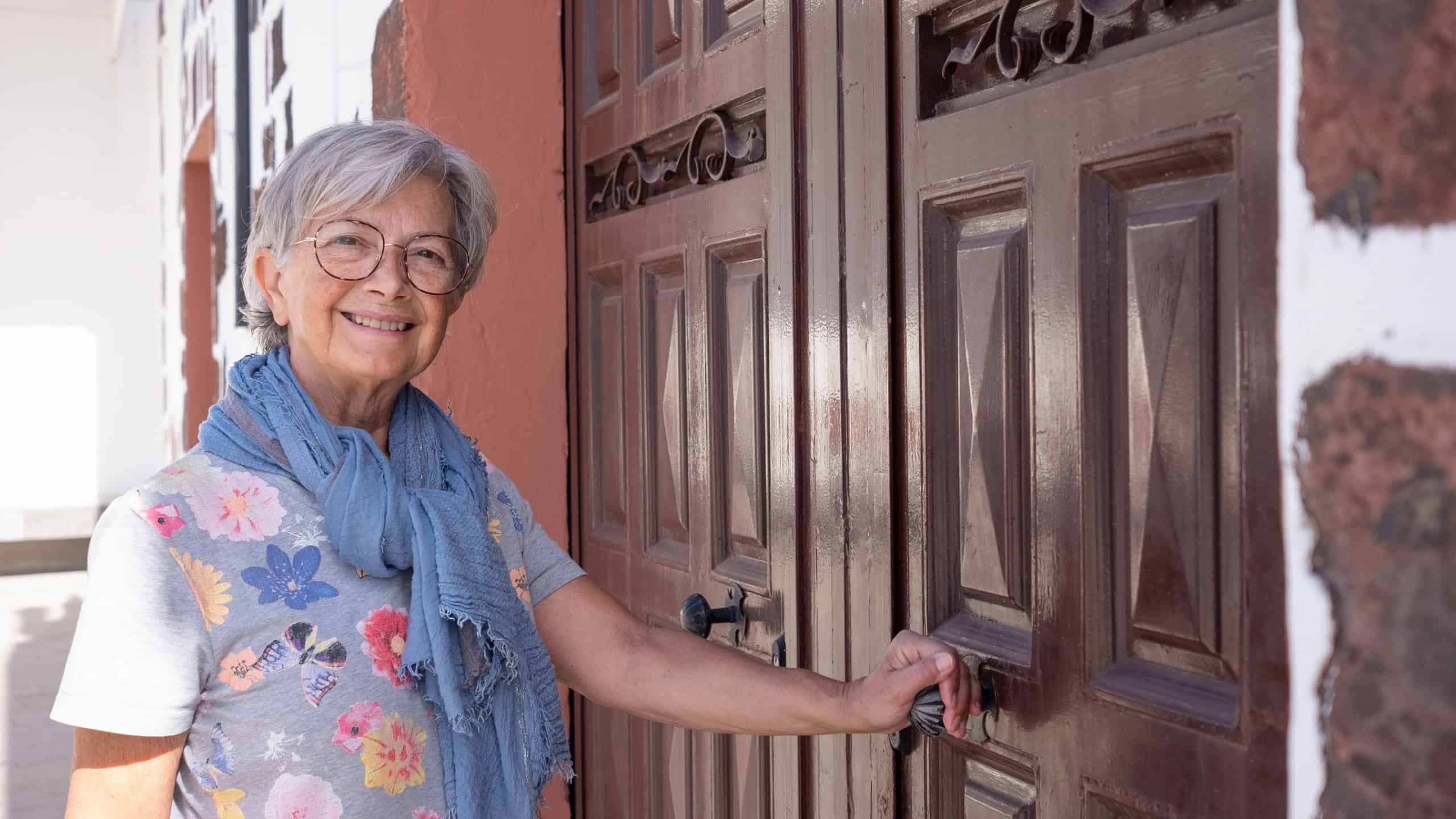 Smiling senior woman short hair wearing eyeglasses and scarf opening a large wooden door of the country house when come back home