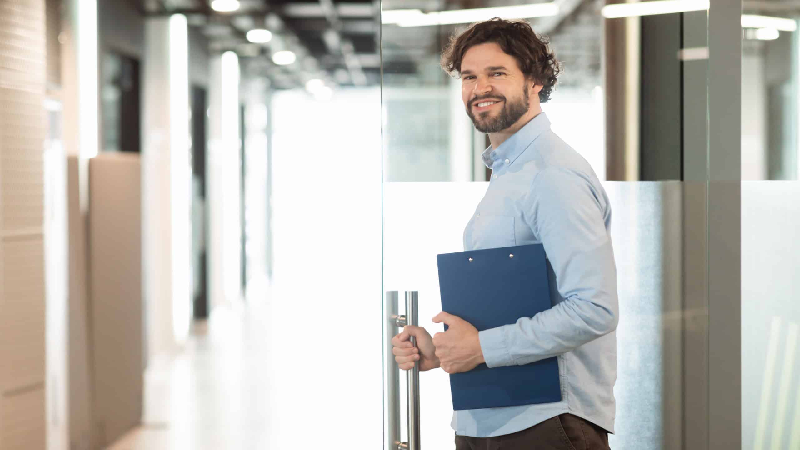 Successful Person. Portrait of confident smiling business man in shirt opening glass door standing at modern office and posing looking back at camera, holding clipboard and handle, free copy space