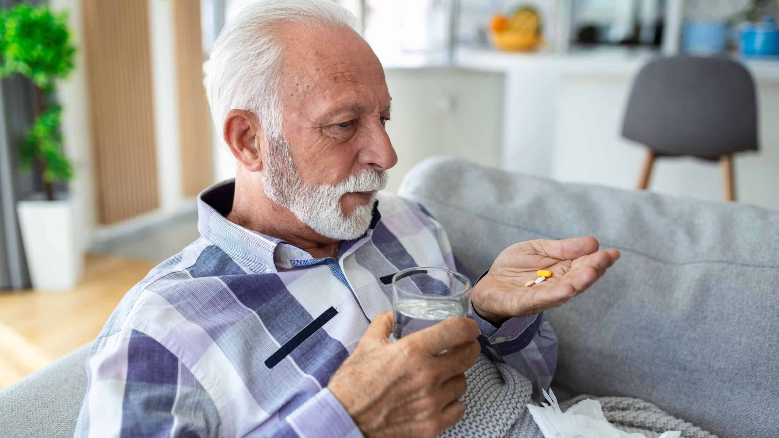 Senior man taking pills while sitting on couch at home, holding white jar with treatment, copy space. Grey-haired elderly man using supplements or vitamins. Healthy lifestyle in senior age concept