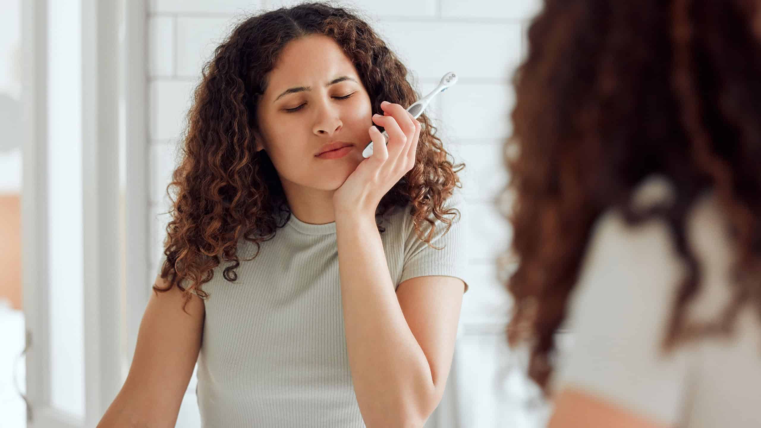 Toothache, oral pain and dental sensitivity for a woman brushing her teeth in the morning. African American female suffering with a painful, hurting or inflammation in her mouth in the bathroom