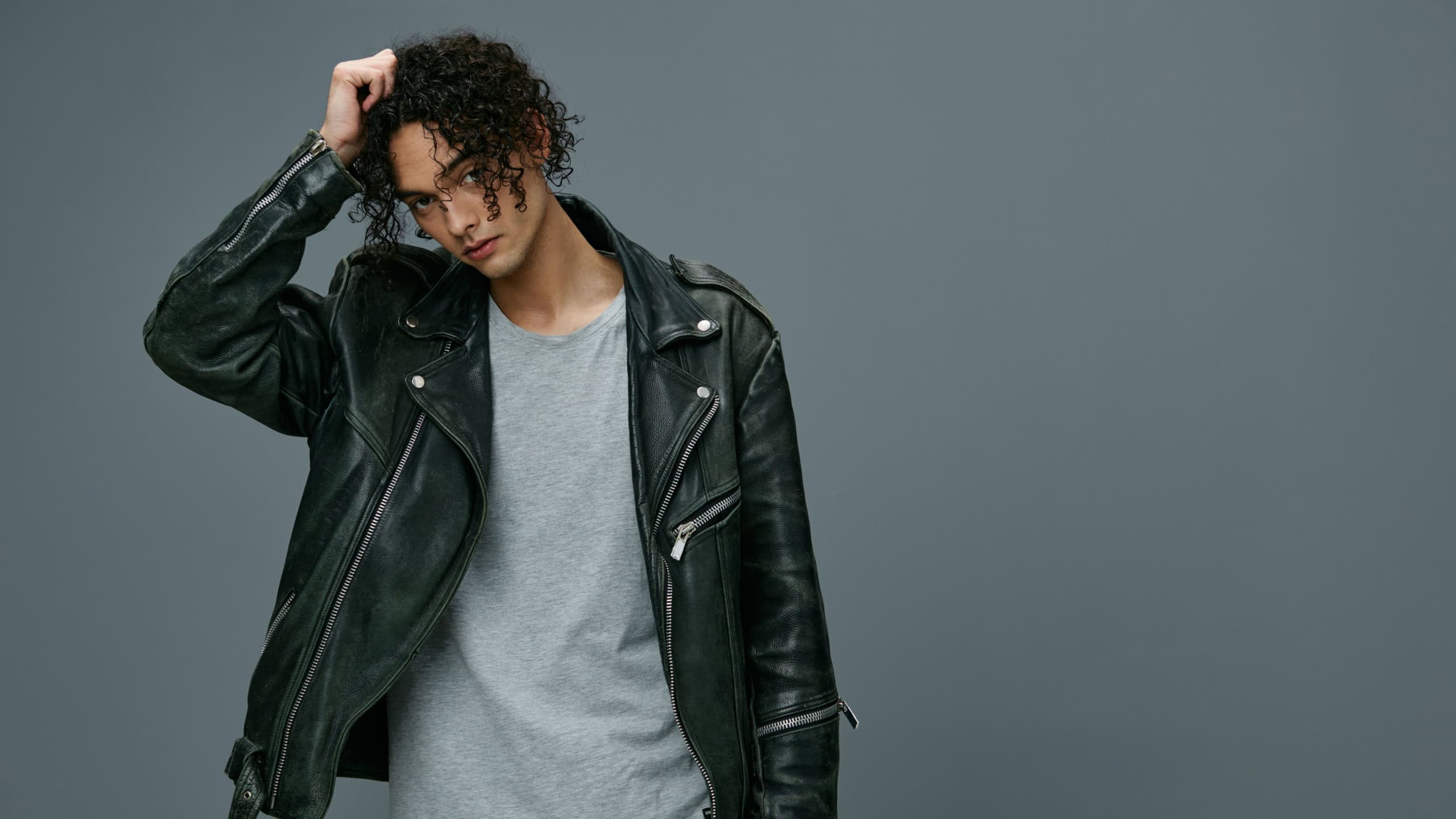 Cool fashion offer. Pretty stylish tanned curly man leather jacket looks at camera posing isolated on over gray studio background. Huge Seasonal Sale New Collection concept. Copy space for ad