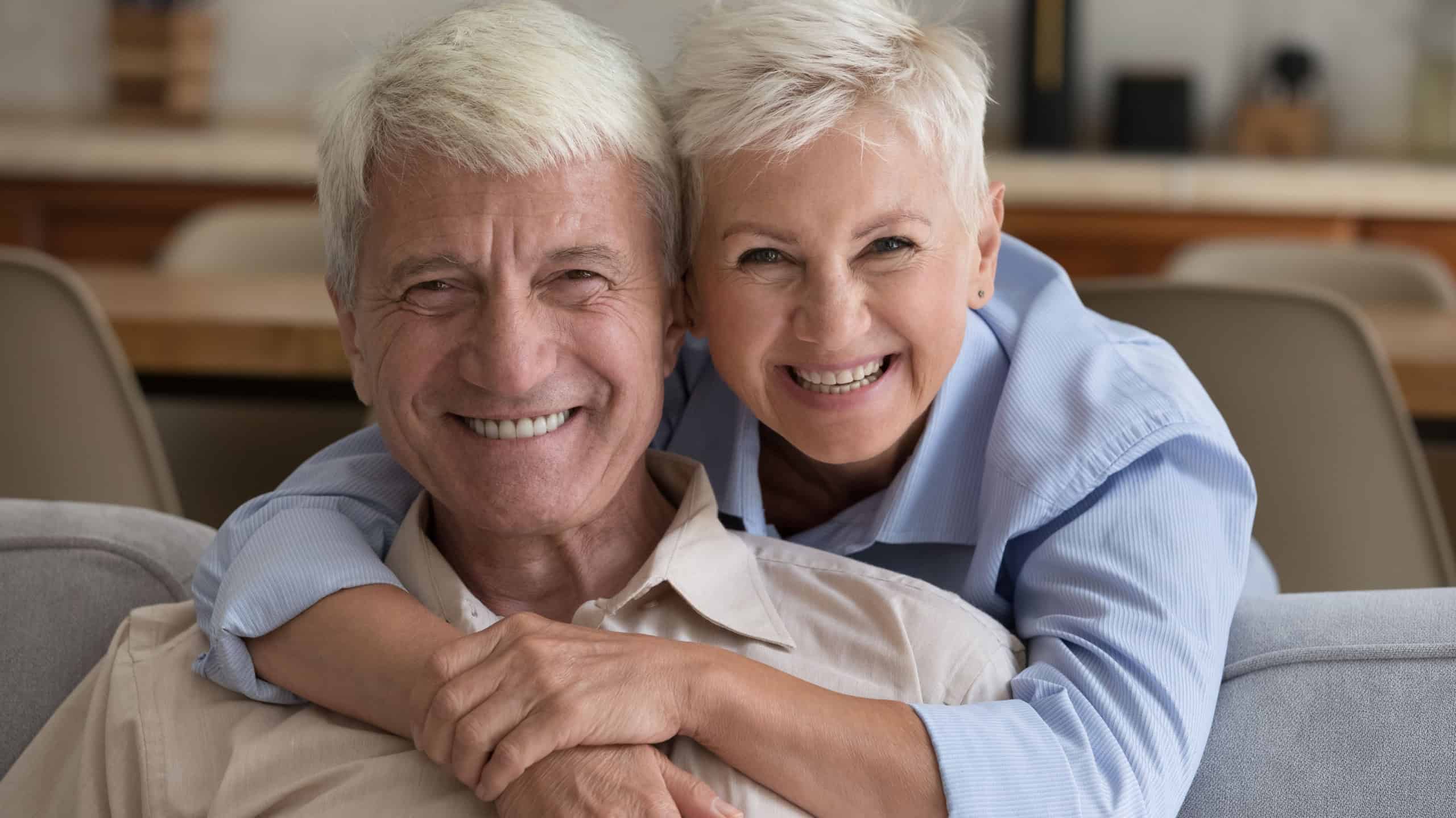 Grey-haired grandparents pose at home. Loving wife hugs husband seated on sofa staring at camera, spend time together. Happy long-lasting marriage, harmonic relations between spouses, respect and bond